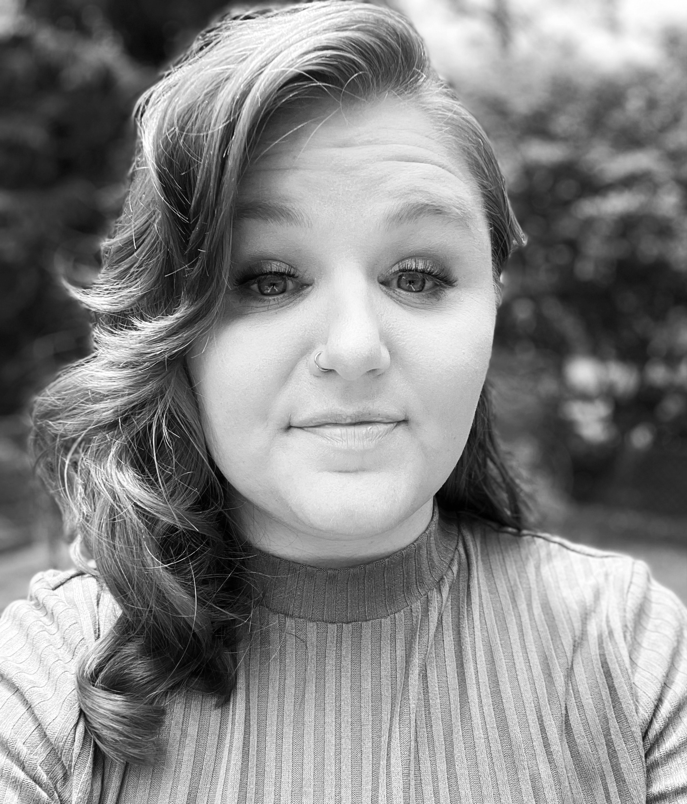 Black and white close-up photo of a woman with curly hair, wearing a striped top and a nose ring, outdoors with blurred background.