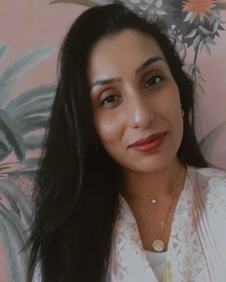 A woman with long black hair, wearing layered necklaces and a white and pastel-colored top, is smiling slightly while looking at the camera. She is standing in front of a floral wall with pastel-colored flowers and leaves.