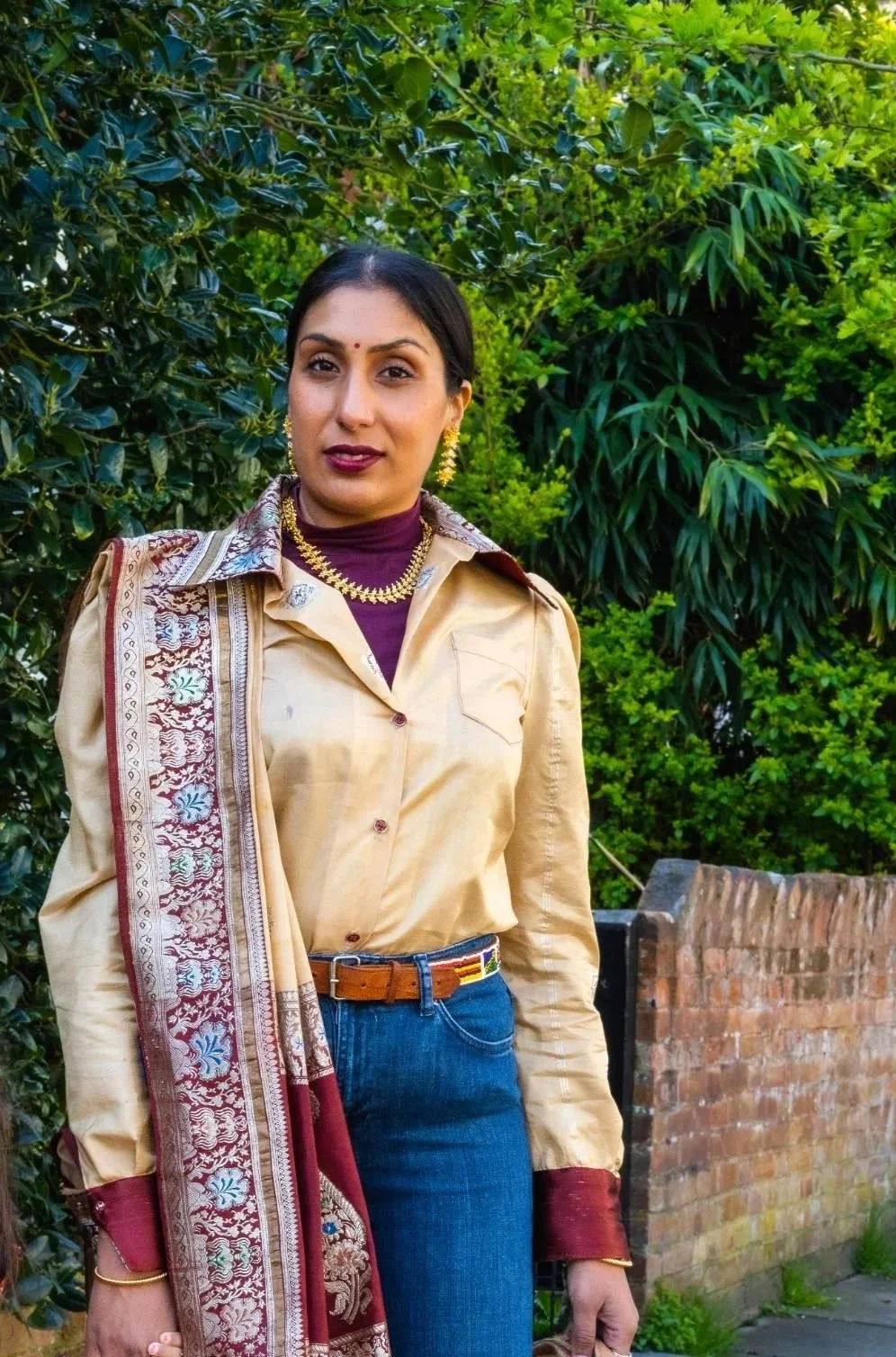 Shilpa standing outdoors in front of green foliage, wearing a upcycled saree shirt in beige with red borders, gold jewellery, and a maroon turtleneck underneath.