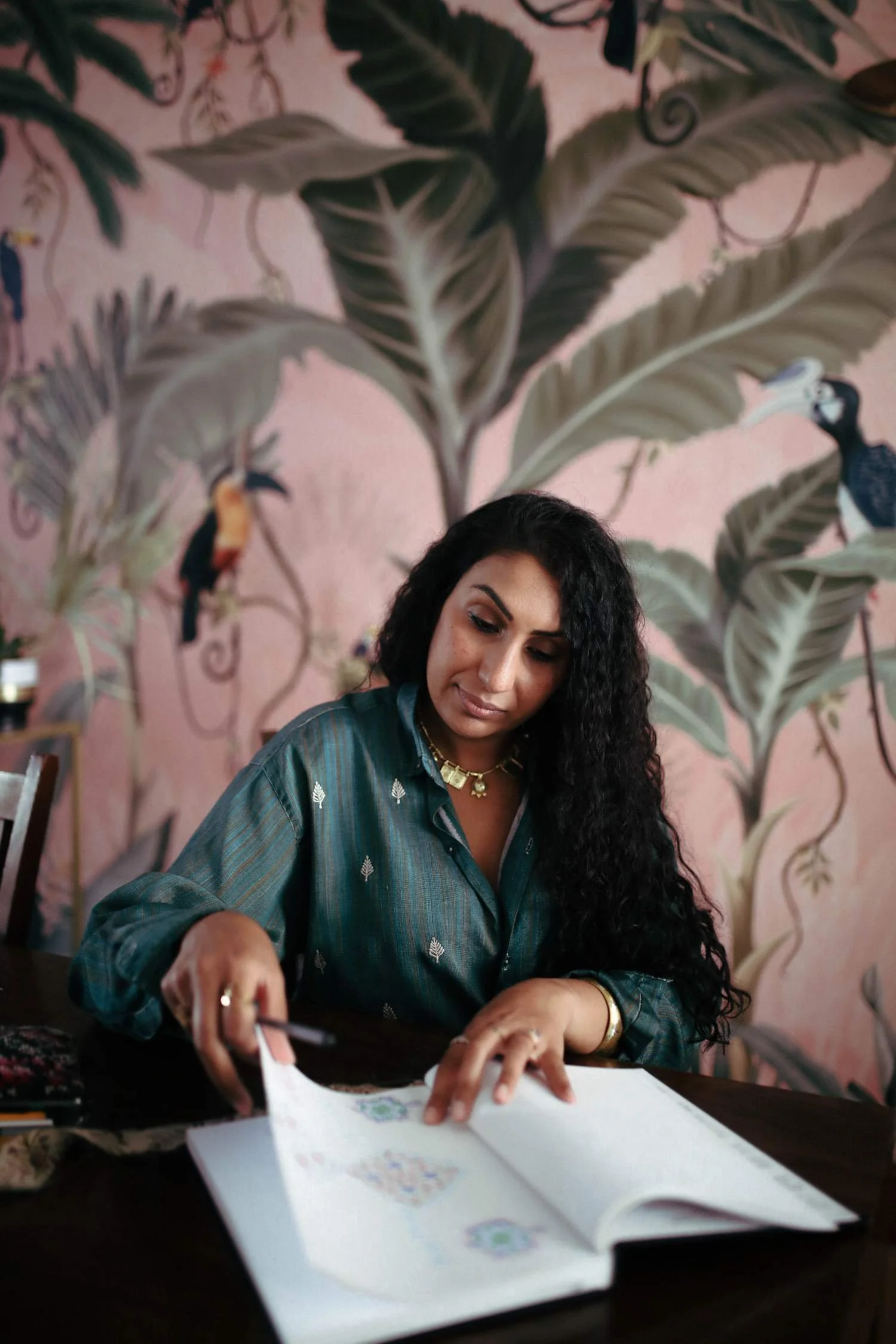 A woman with long, curly black hair looking down at an open book with colorful illustrations. She is wearing a teal shirt with small white patterns and gold jewelry, seated at a dark wooden table. The background features a pink wall with large, painted green leaves and colorful bird illustrations.