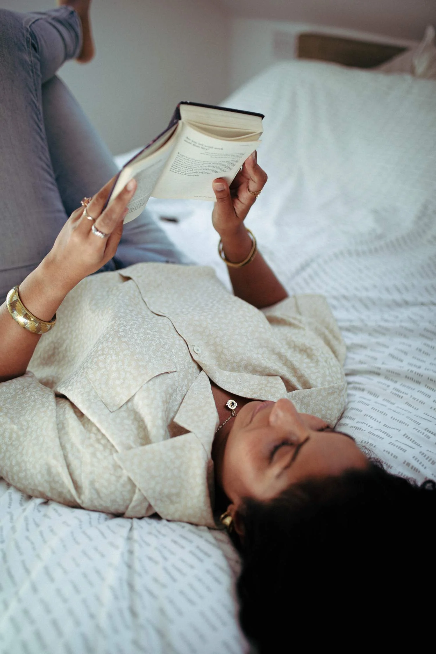 Shilpa lying on her back on a bed, holding and reading a book, with her head resting on a pillow and her eyes closed.
