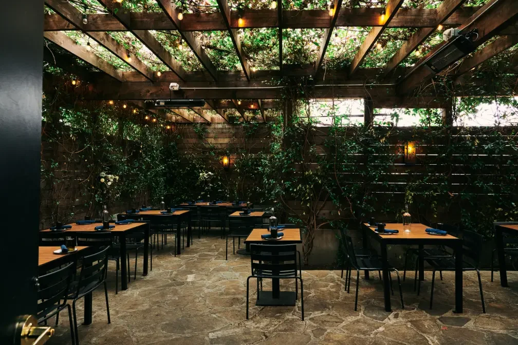 A cozy outdoor dining area with wooden tables and black chairs, decorated with dark napkins and water bottles, surrounded by green plants and string lights hanging from a wooden pergola.