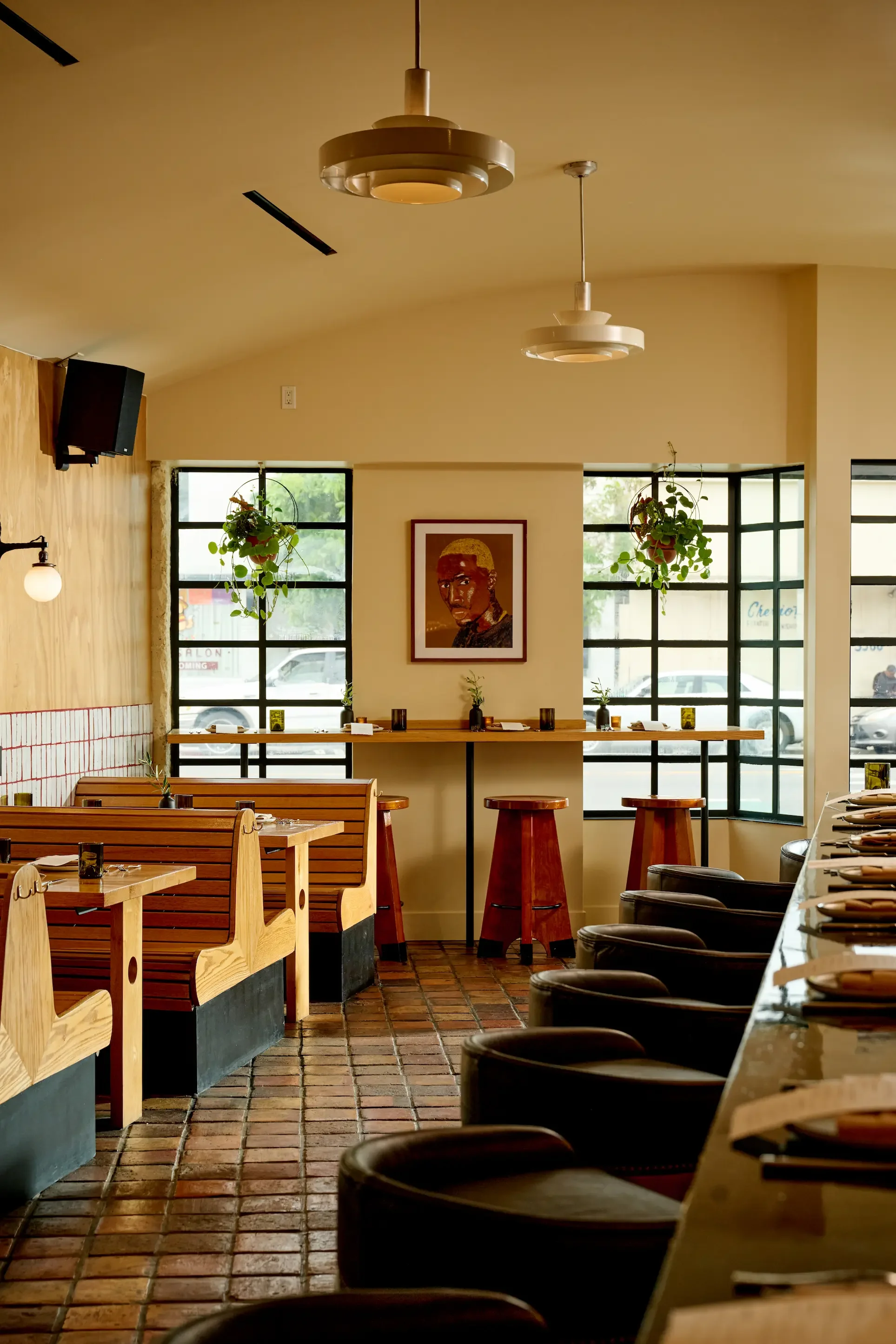 Interior of a cozy restaurant with wooden tables and black chairs, hanging plants, framed artwork, windows with natural light, and pendant lighting.