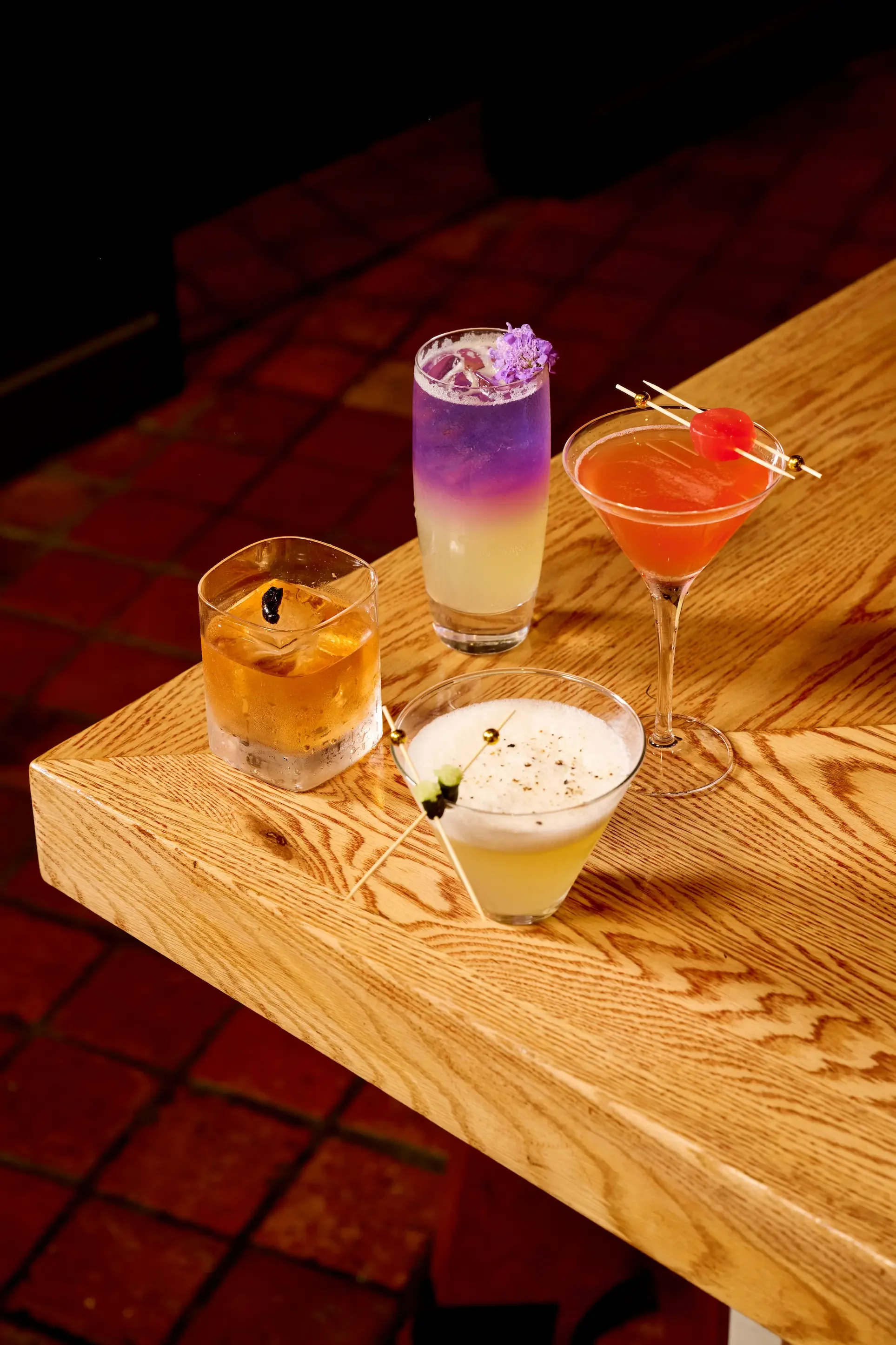 Assorted cocktails on a wooden bar counter with brick flooring in the background.