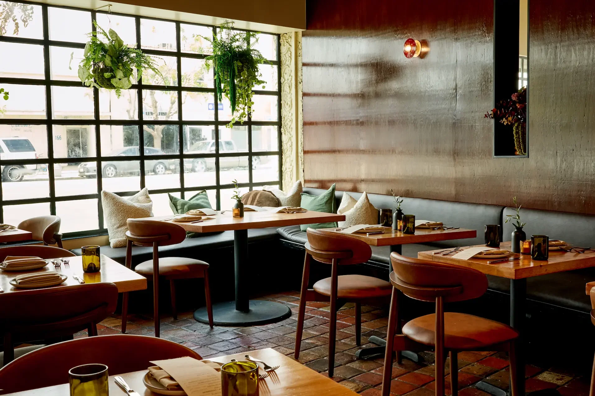 Interior of a restaurant with wooden tables set with plates, silverware, and glasses, decorated with small vases of greenery, and a large window with hanging plants, warm lighting, and a textured wall with built-in floral display.