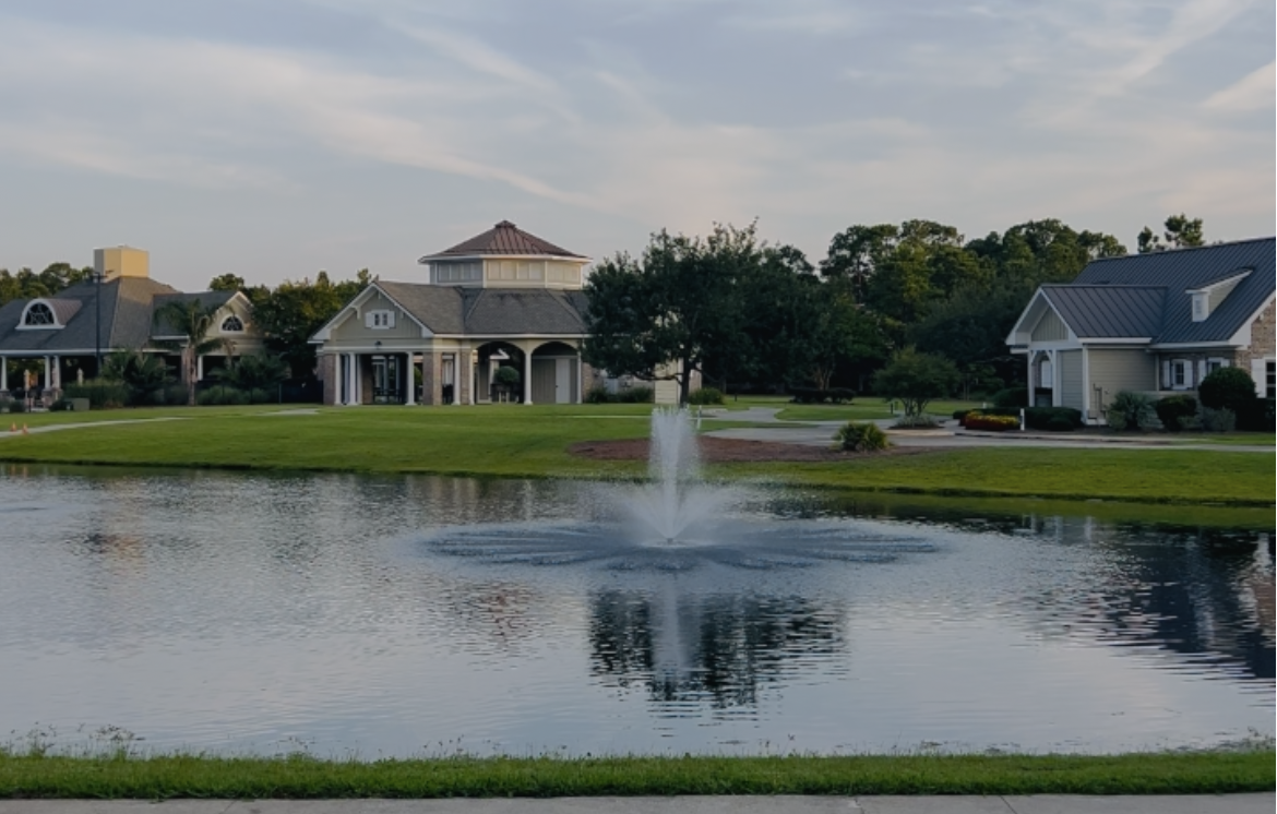 A pond with a fountain in front of a row of residential houses with well-maintained lawns and trees under a partly cloudy sky.