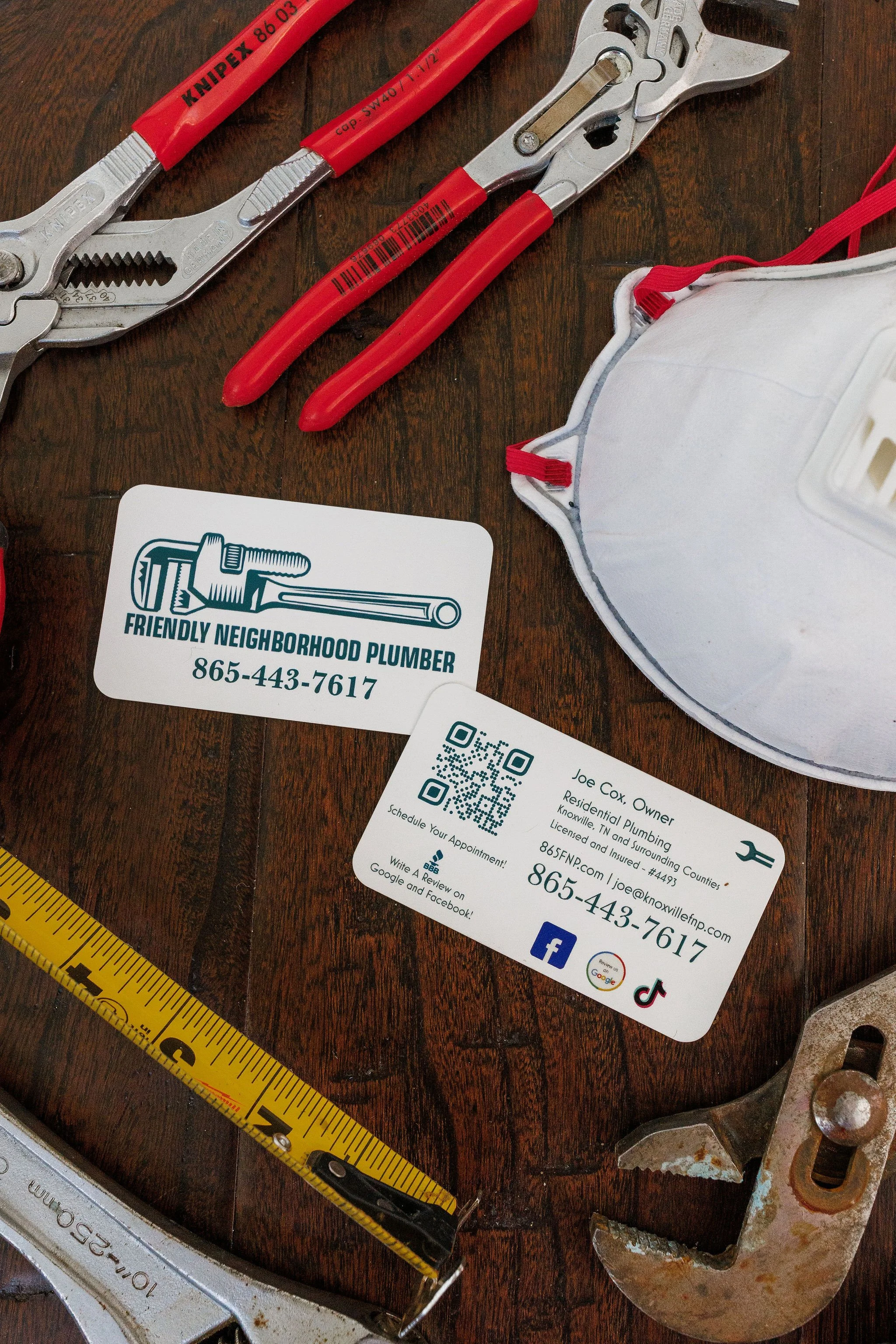 Business cards for a plumbing company, plumber's wrench, pliers, a tape measure, and a part of a rusty pipe or fitting on a wooden surface.