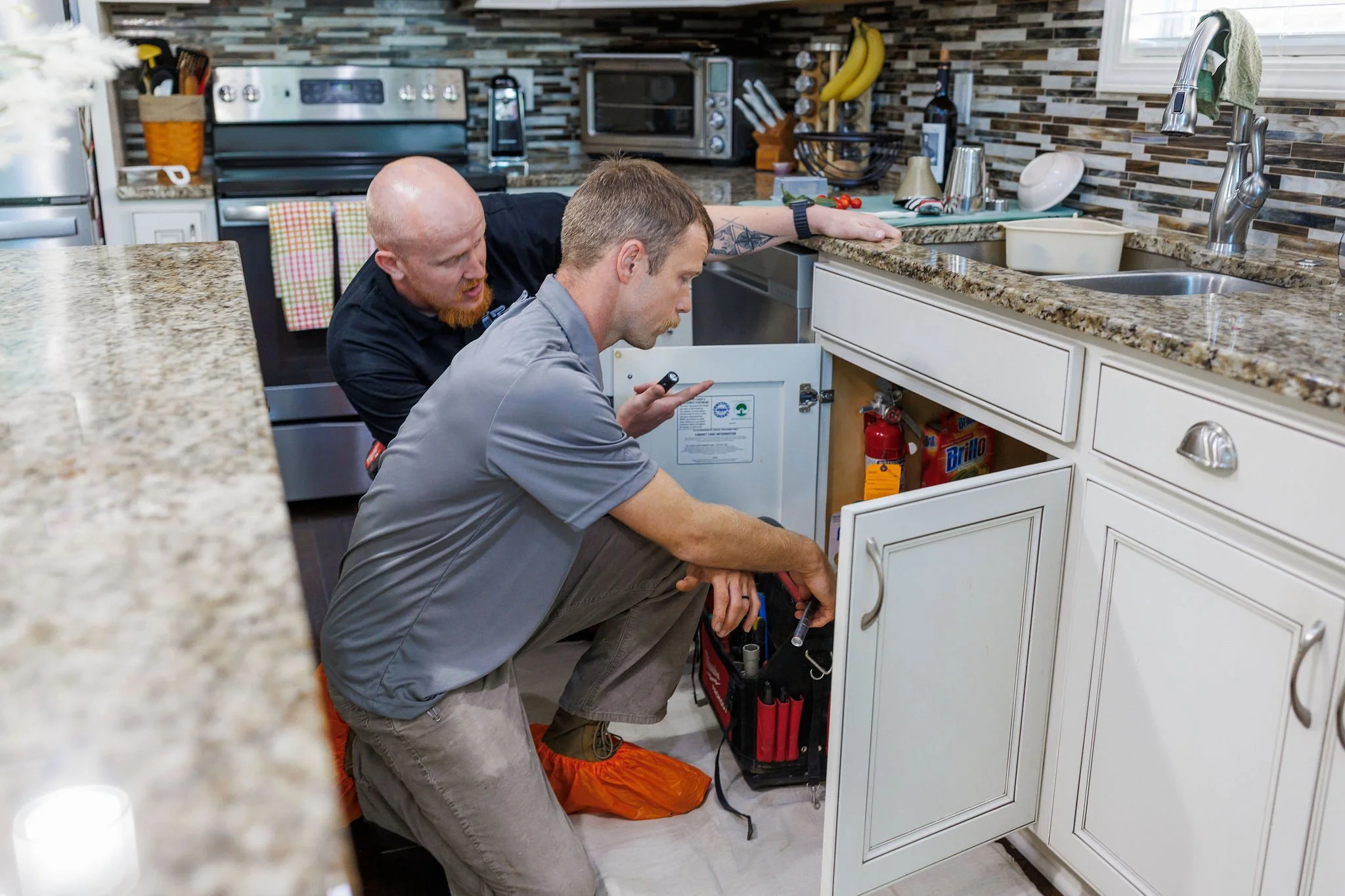 Two men working on kitchen plumbing under the sink with kitchen appliances and utensils visible in the background.