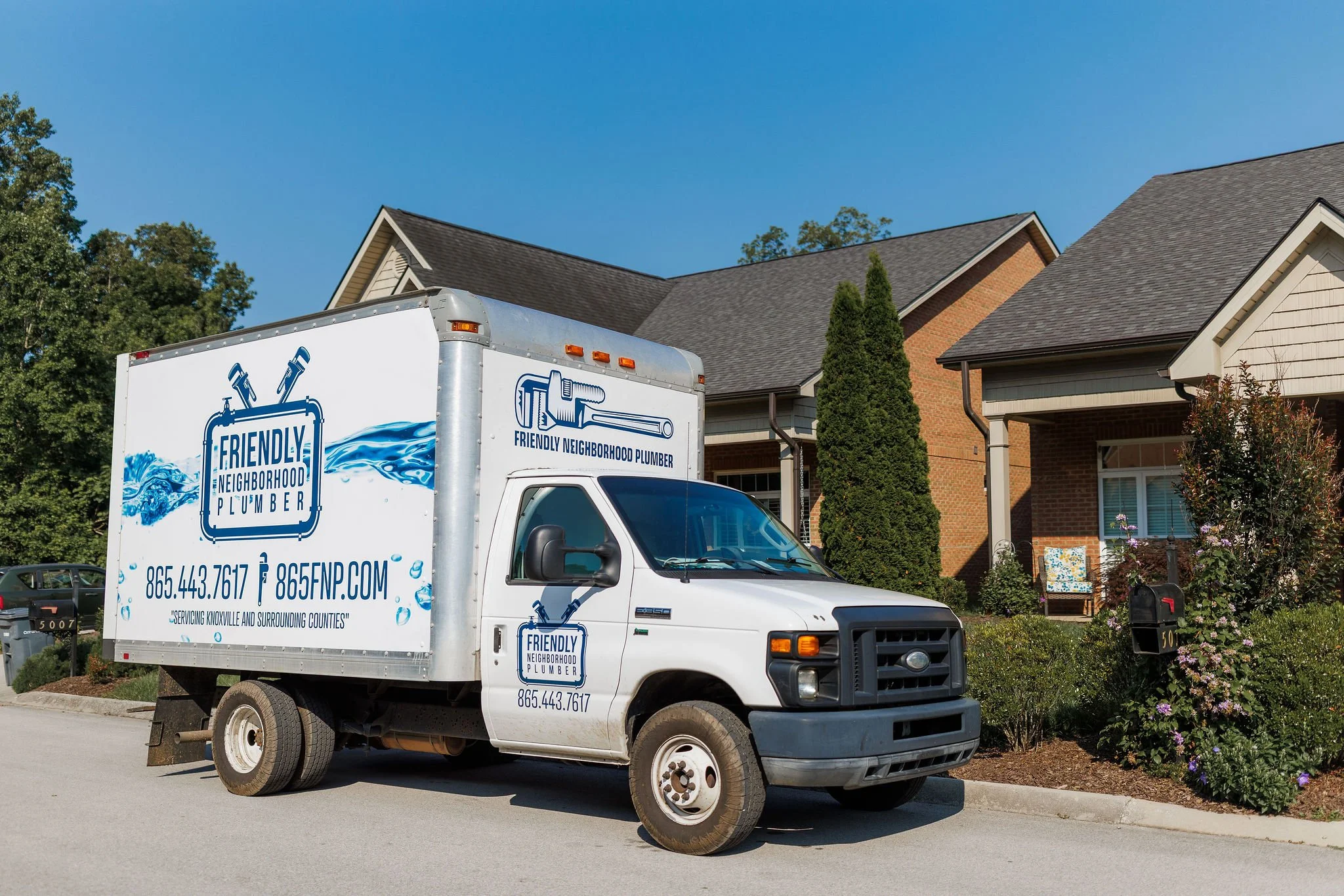 A white plumber's service truck parked in front of a residential house with gray roofs and brick siding. The truck has the company name 'Friendly Neighborhood Plumber' and contact information on the side.