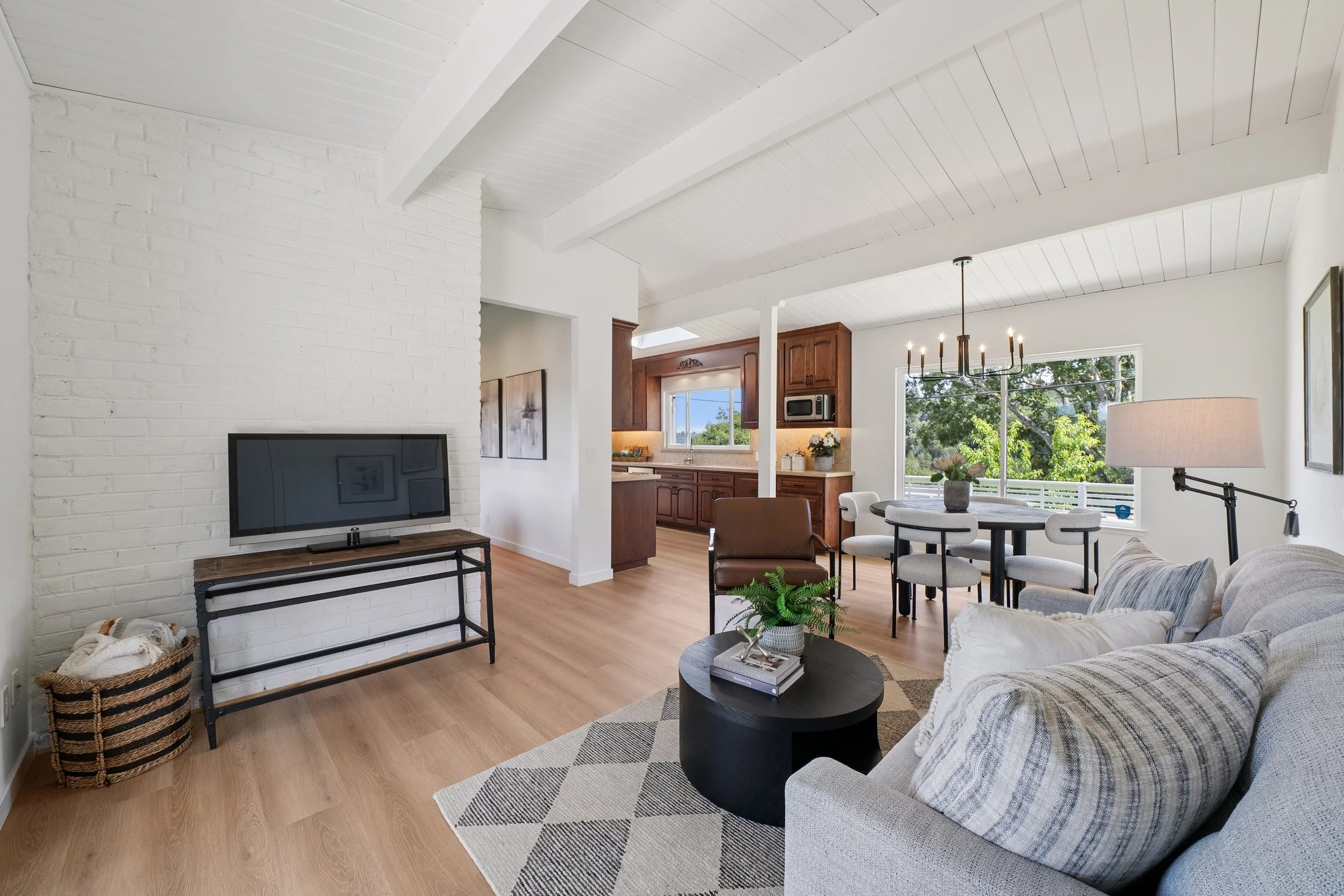 Living room with white brick wall, TV on a stand, wooden flooring, beige sofa with pillows, round black coffee table with books and plant, dining area with round table, white chairs, chandelier, open kitchen with wooden cabinets, window showing trees outside.