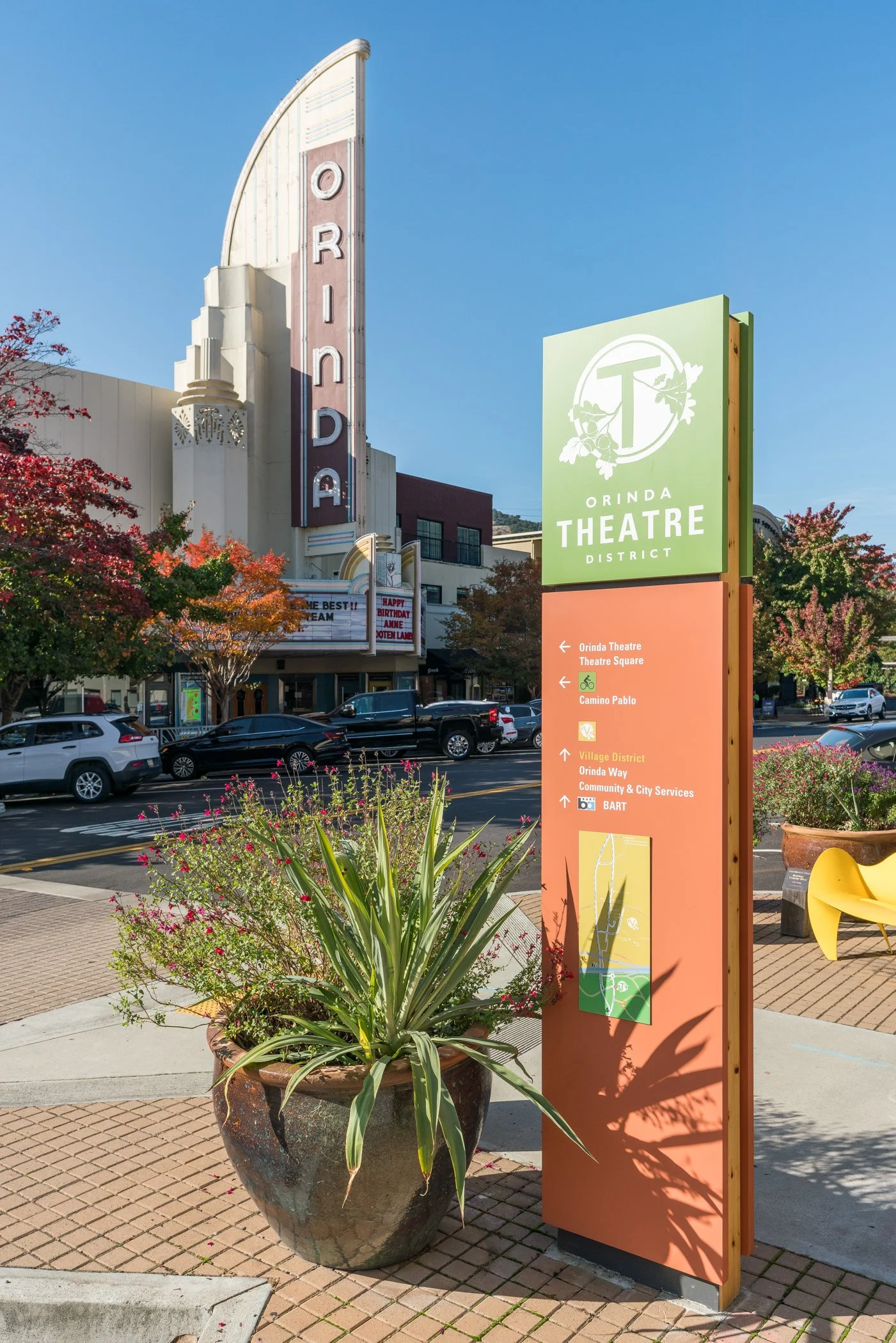 Colorful downtown street scene in Orinda with a large theater sign, trees with fall foliage, parked cars, and a bright blue sky.