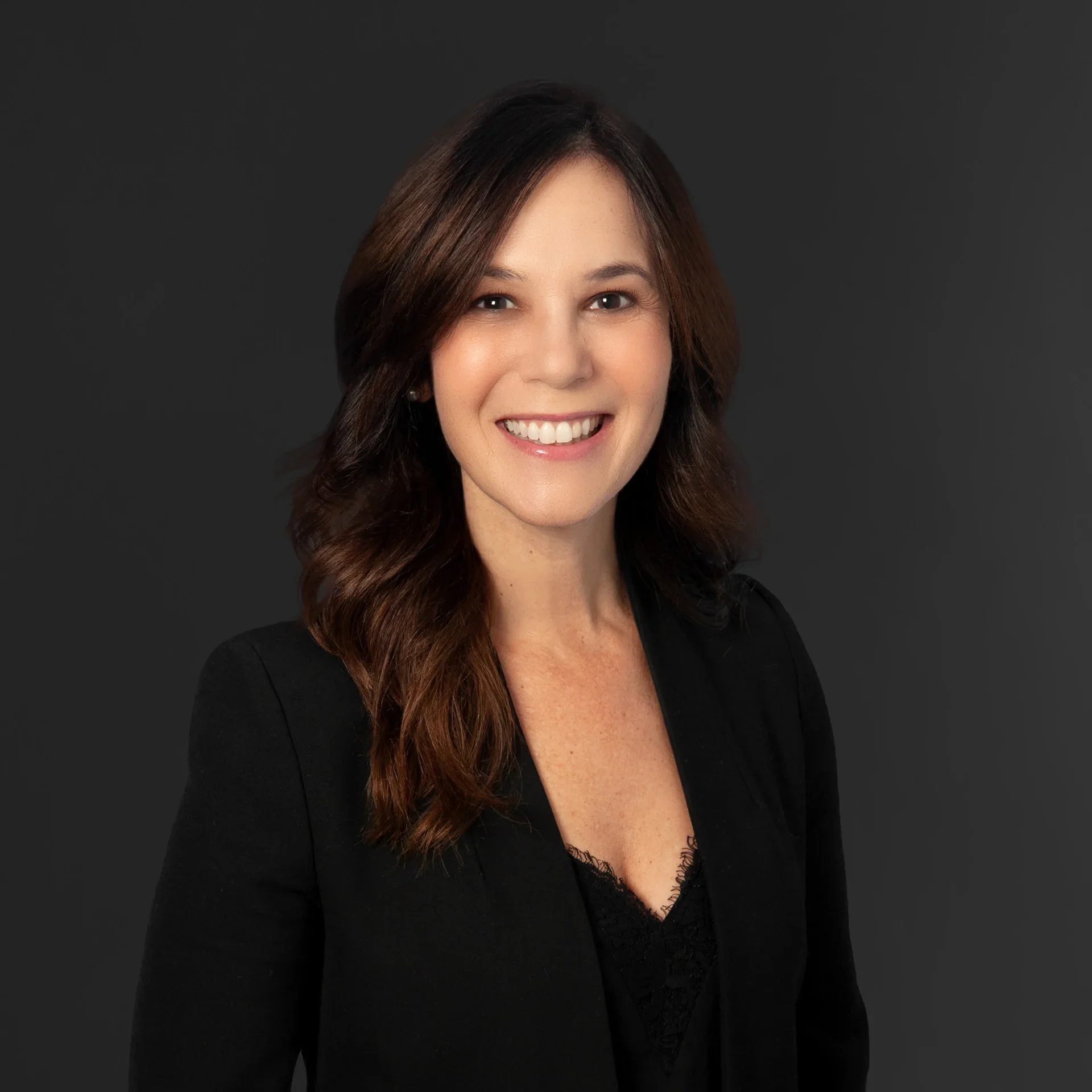 Professional woman with long wavy brown hair, wearing a black blazer and smiling, against a dark gray background.