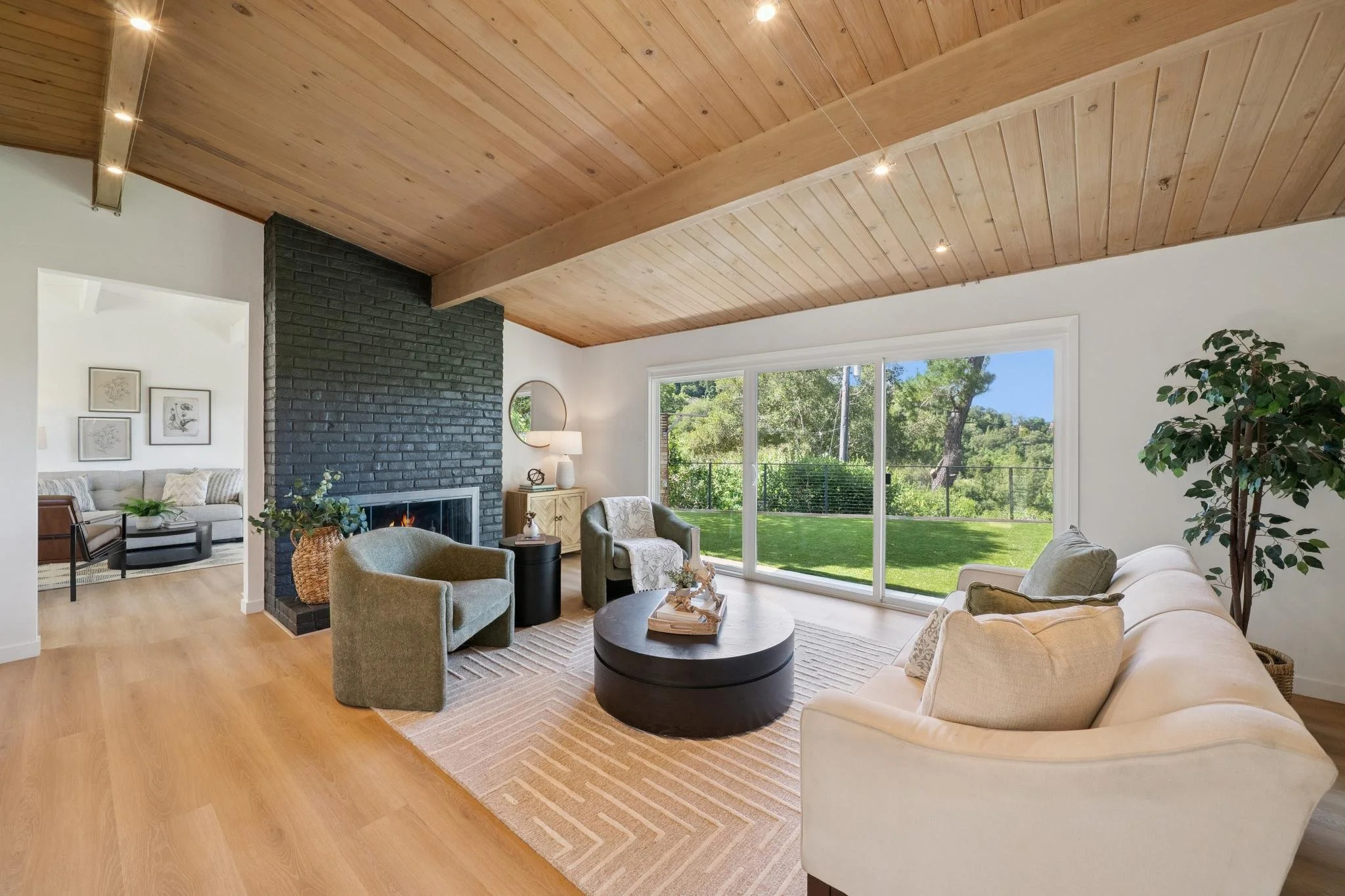 Bright living room featuring a beige couch, green armchairs, a black round coffee table, and a wooden ceiling with spotlights. Large sliding glass doors open to a lush green yard with trees.