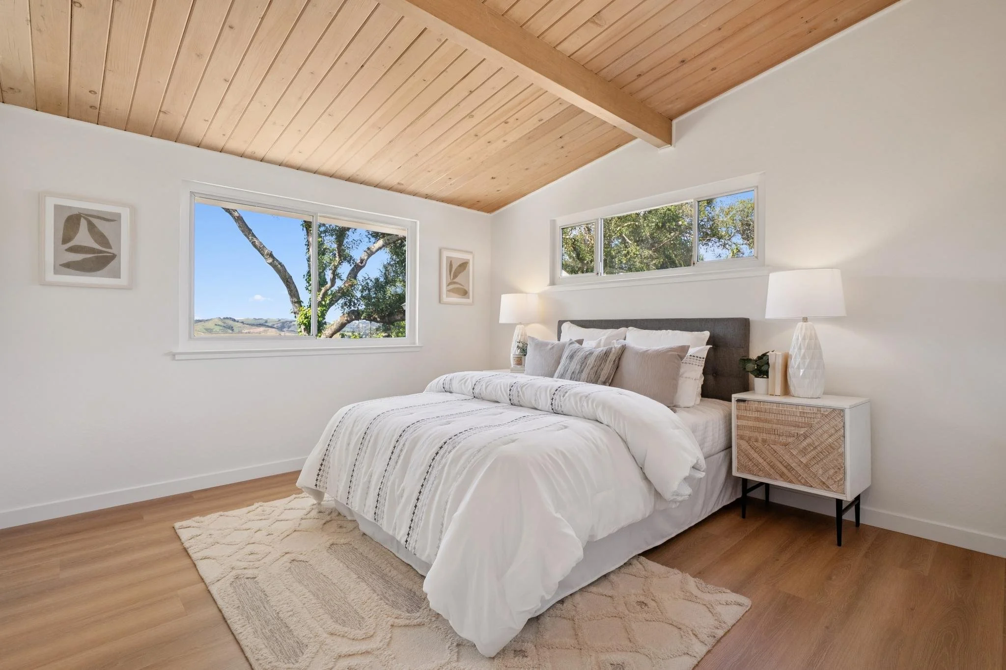 A modern bedroom with a wooden ceiling, white walls, a large bed with white and beige bedding, and a nightstand with a white lamp and a small plant. There are two windows showing green trees and a landscape outside.