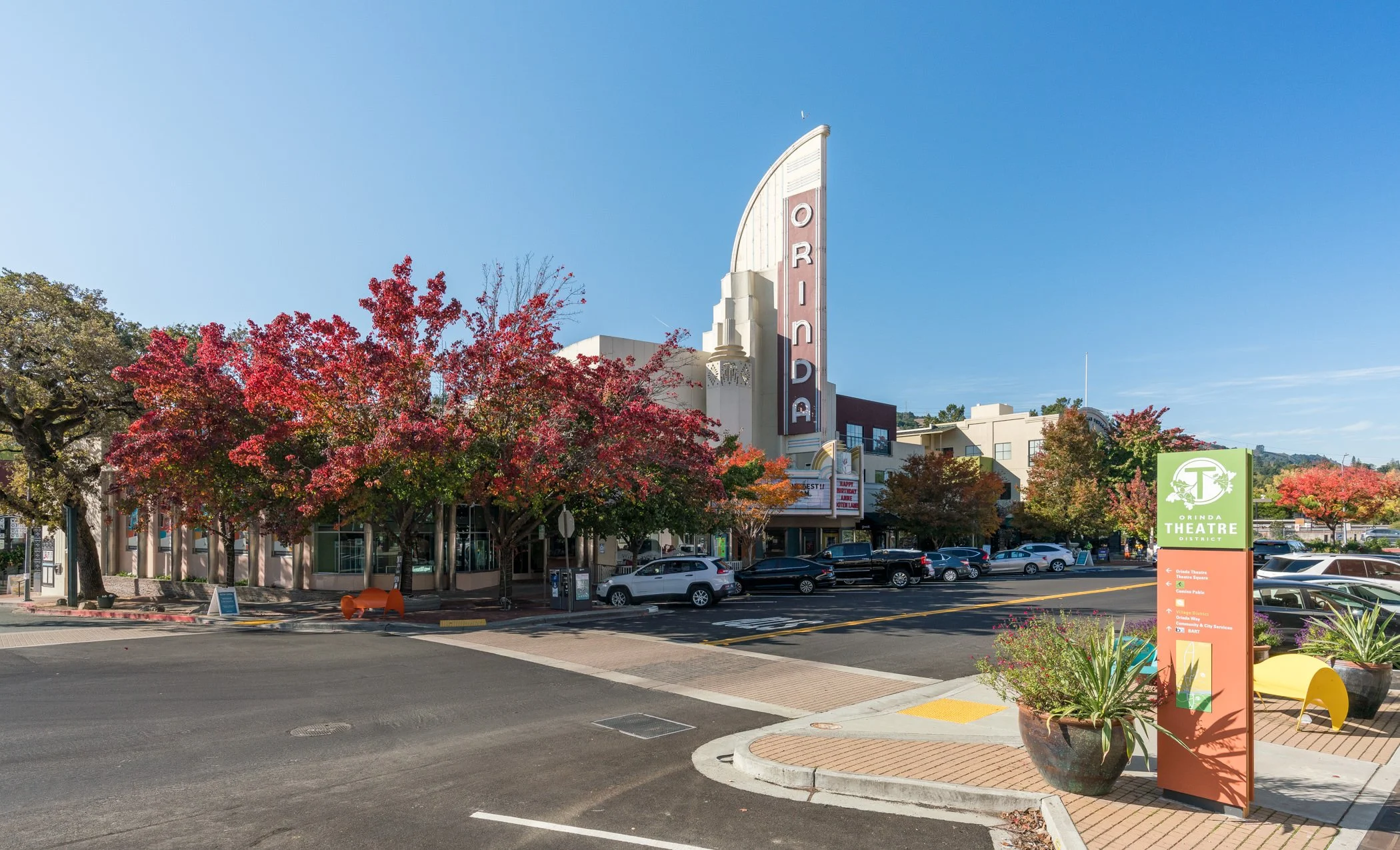 Street view of Orinda Theatre with colorful fall trees, parked cars, and a green district sign.