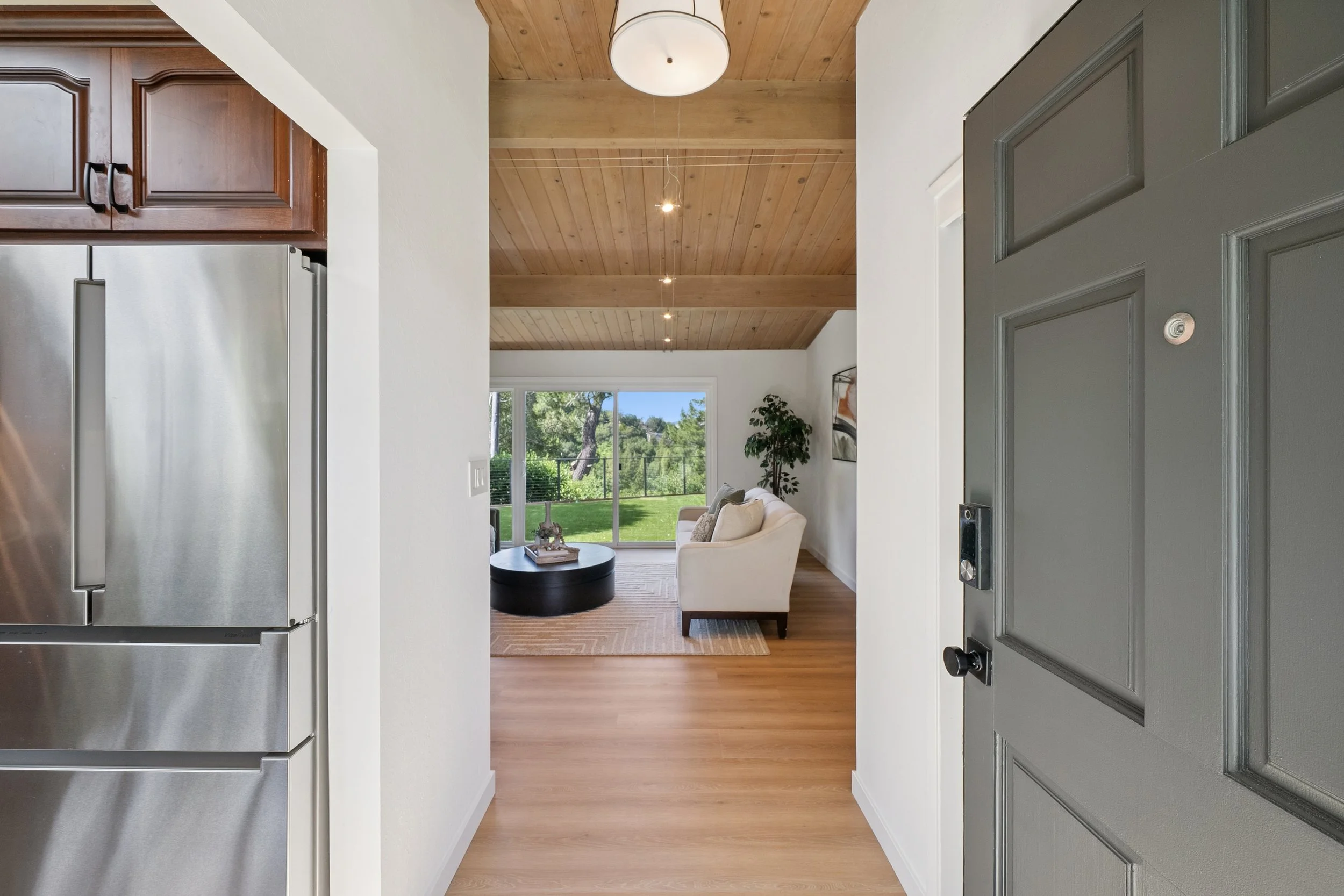 View from the doorway of a living room with a sloped wooden ceiling, white walls, a large window, beige sofa, black coffee table, and green outdoor scenery.