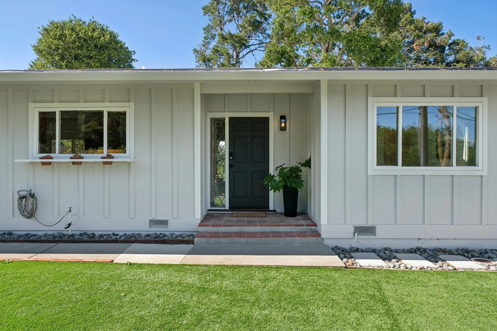 Front of a house with white siding, a dark green front door, and two large windows, one on each side of the door. There is a small porch with brick steps, a potted plant next to the door, and a green lawn in the foreground. Trees can be seen in the b