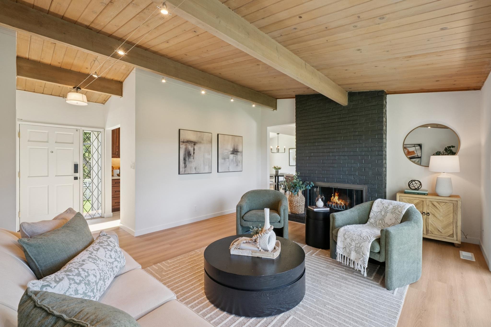 Living room with wood ceiling, black brick fireplace, beige sofa with throw pillows, green armchairs, round black coffee table, wall art, window door, side table with lamp, decorative items