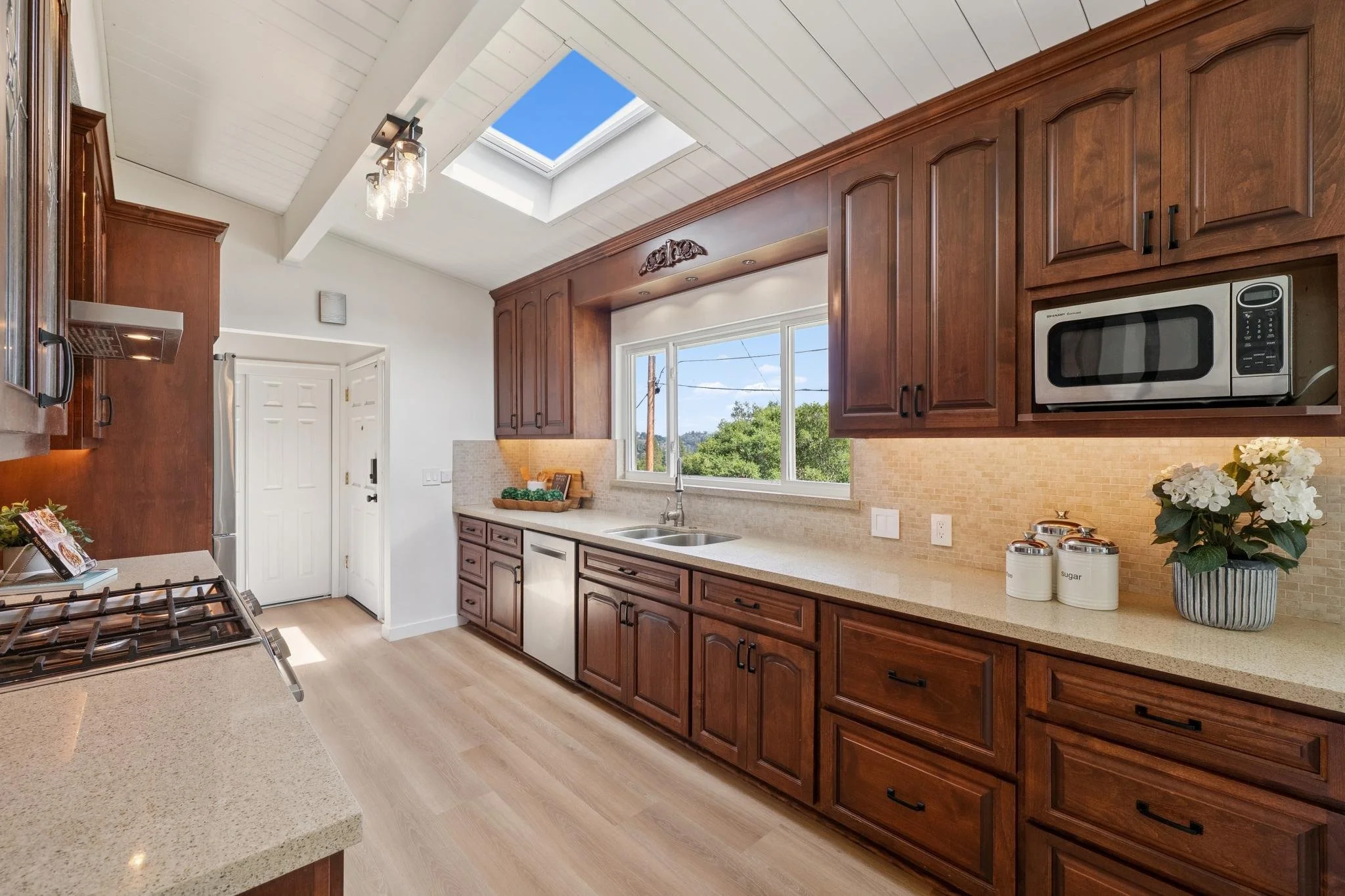 A well-lit kitchen with brown wooden cabinets, a large window above the sink, a skylight, and beige countertops. There's a microwave, a potted plant, and containers labeled sugar and flour on the counter.