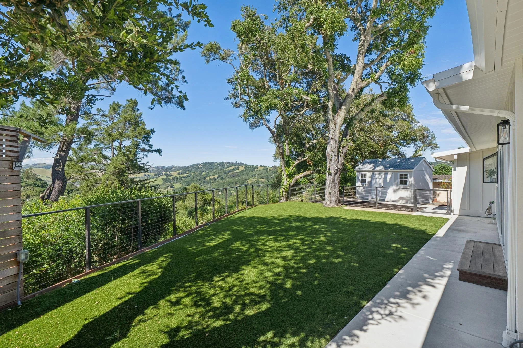 Beautiful backyard with green grass, large trees, and a view of rolling hills in the distance. The yard is bordered by a black metal fence, with a house on the right side and a small shed or outbuilding further back.