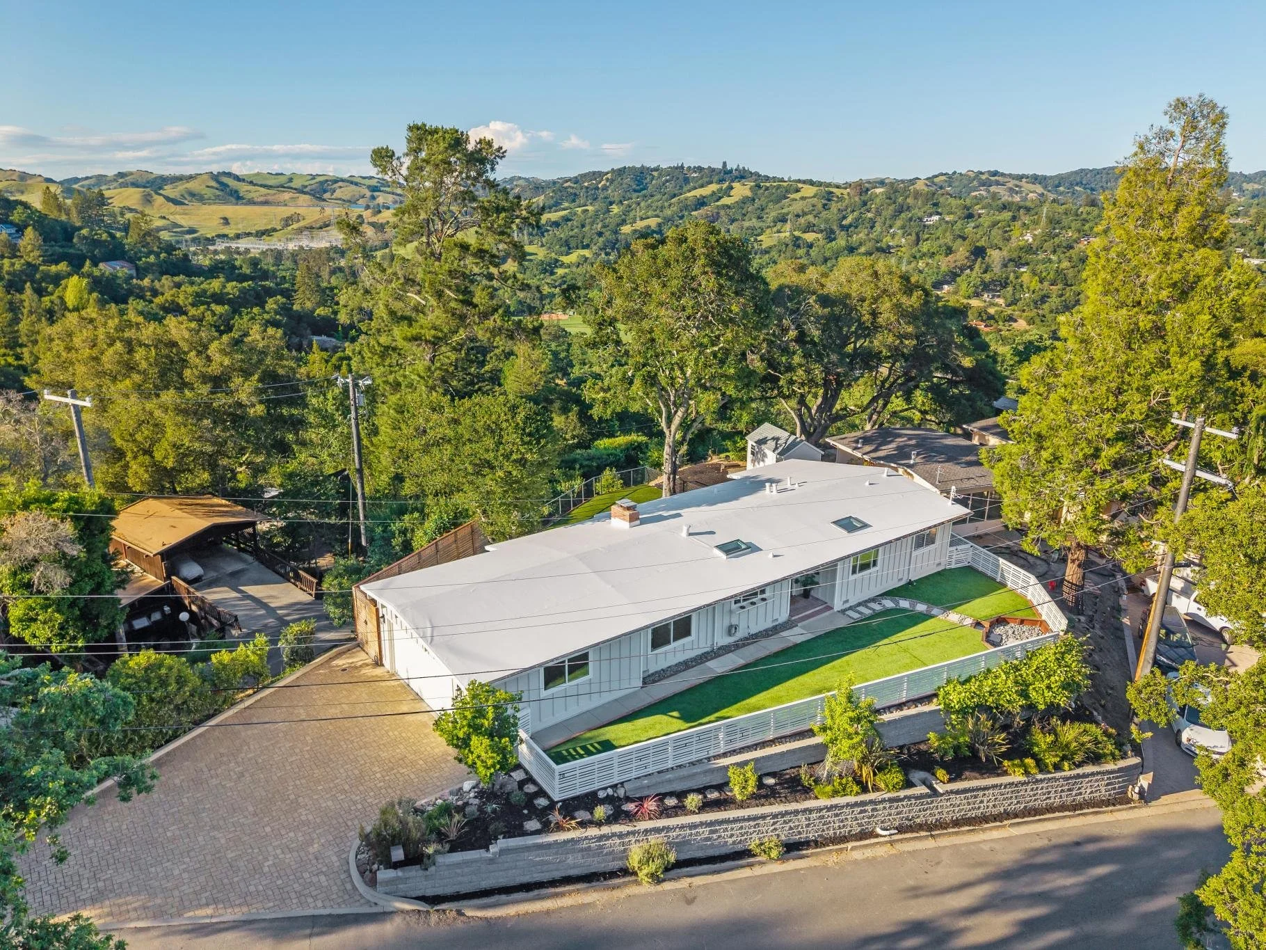  aerial view of a modern white house with a large roof, surrounded by a white fence, trees, green lawn, and neighboring houses in a hilly landscape under a clear blue sky.