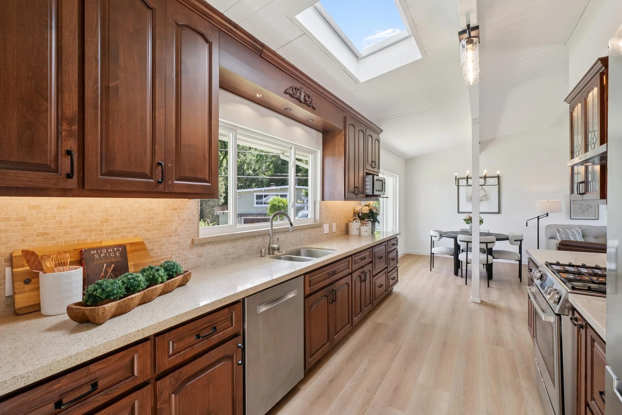 Bright kitchen with wooden cabinets, beige countertops, stainless steel appliances, large window, and a skylight. Open dining and living area with light-colored furniture.