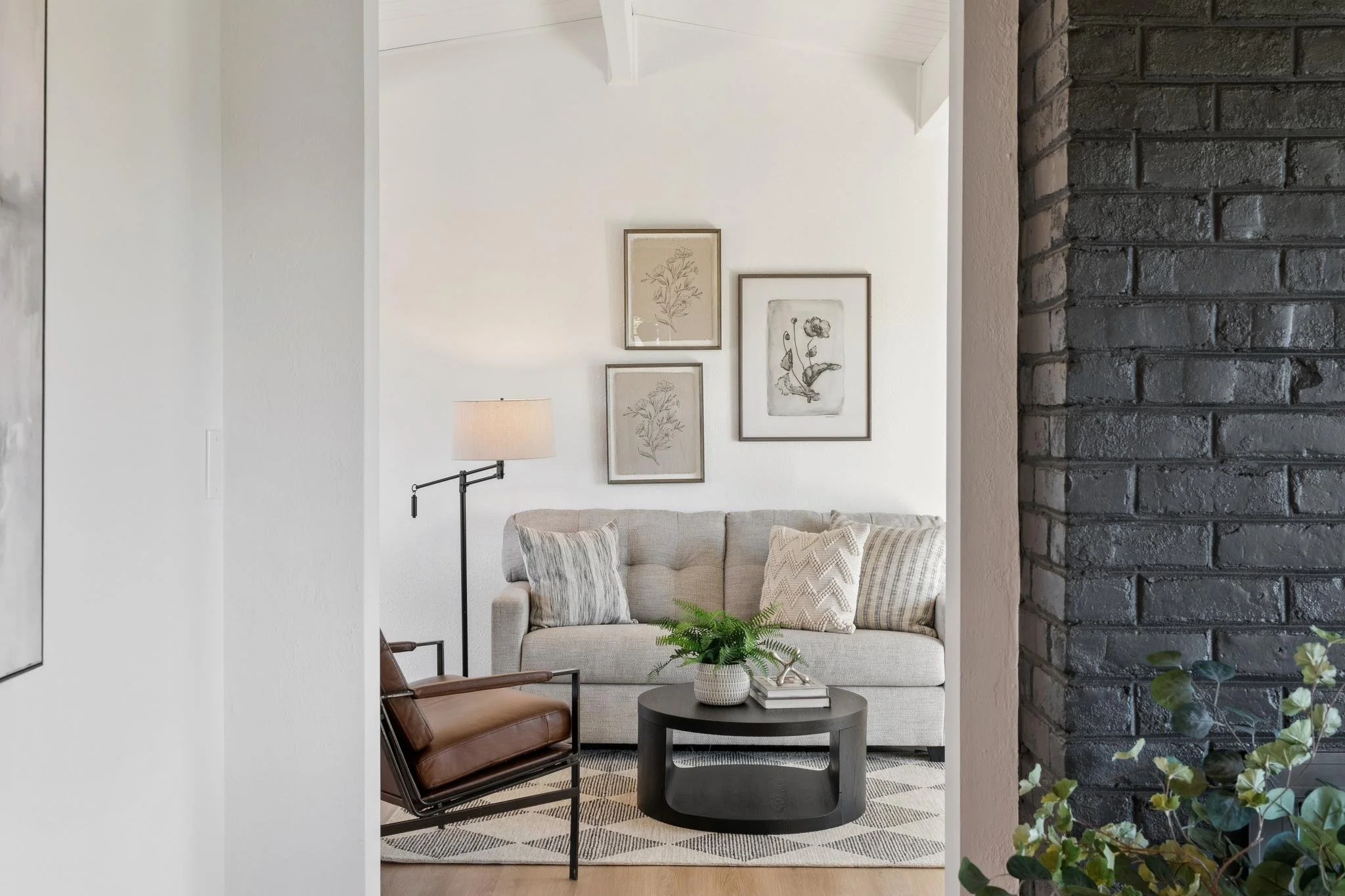 Living room with beige sofa decorated with patterned pillows, black coffee table with a potted plant and books, art prints on the white wall, a brown leather chair, and a wall lamp, with a black brick wall on the right side.