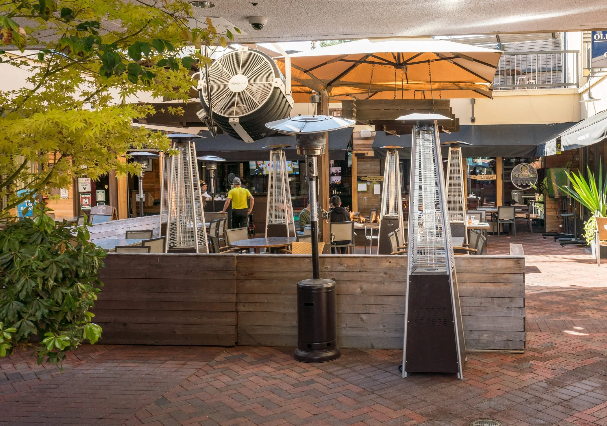 Outdoor dining area with patio heaters and a fire pit, enclosed by a wooden fence, in front of a restaurant with patrons and staff visible.