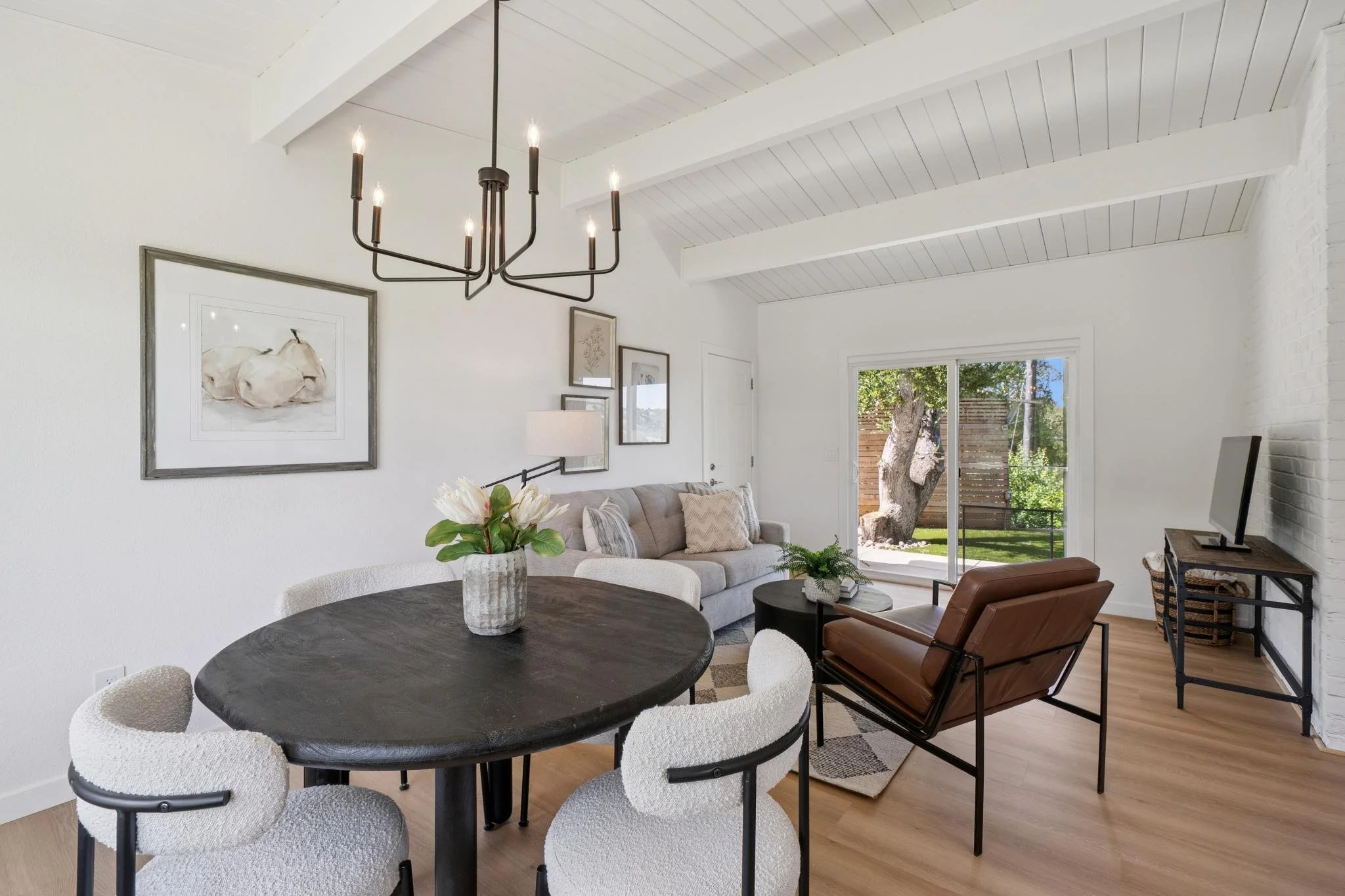 Living room with round dining table, white upholstered chairs, beige sofa, leather chair, and sliding glass door to outdoor yard with tree.