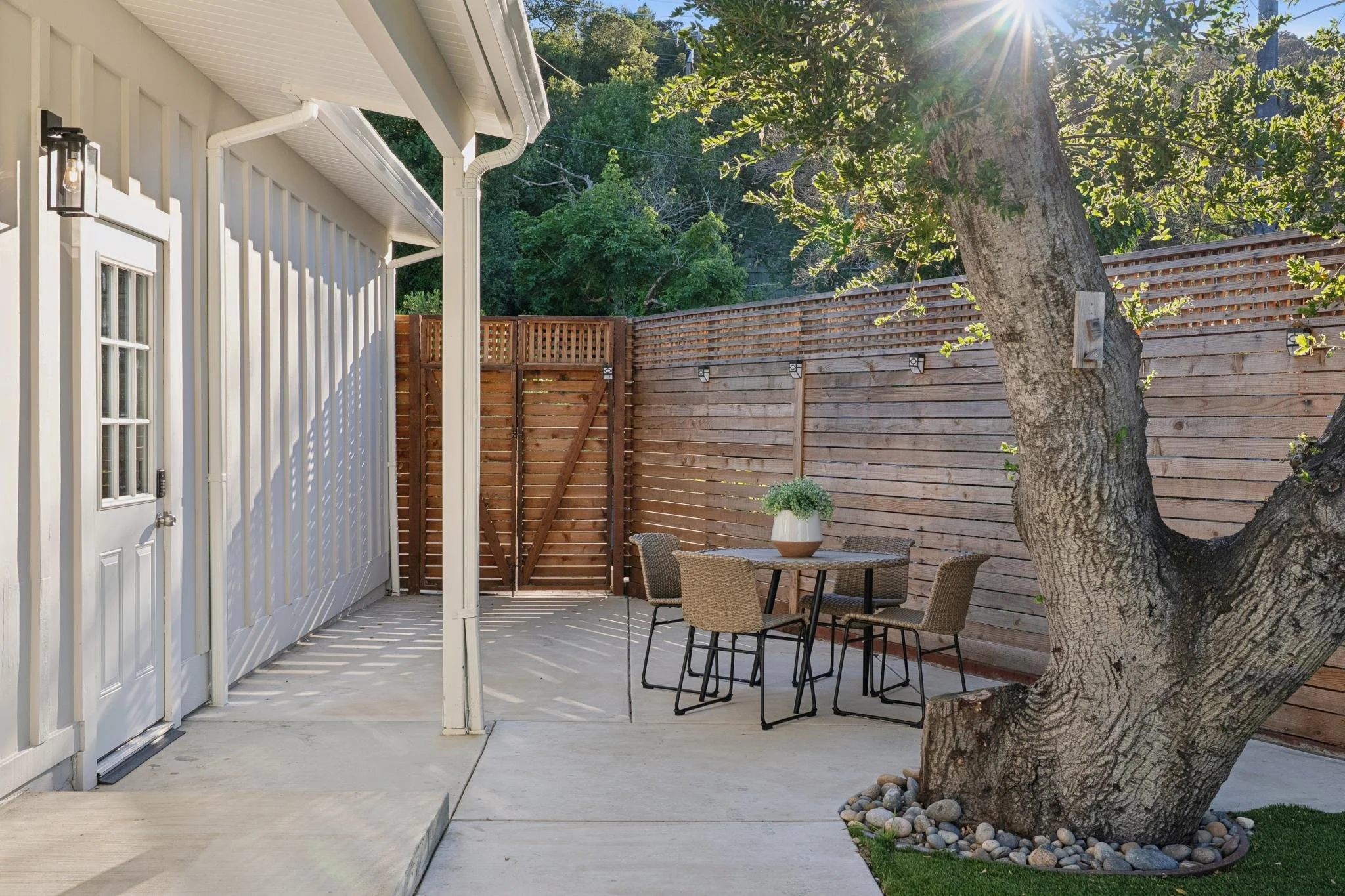 A cozy backyard patio with a wooden fence, a mature tree, an outdoor table with four chairs, and a potted plant, bathed in sunlight.
