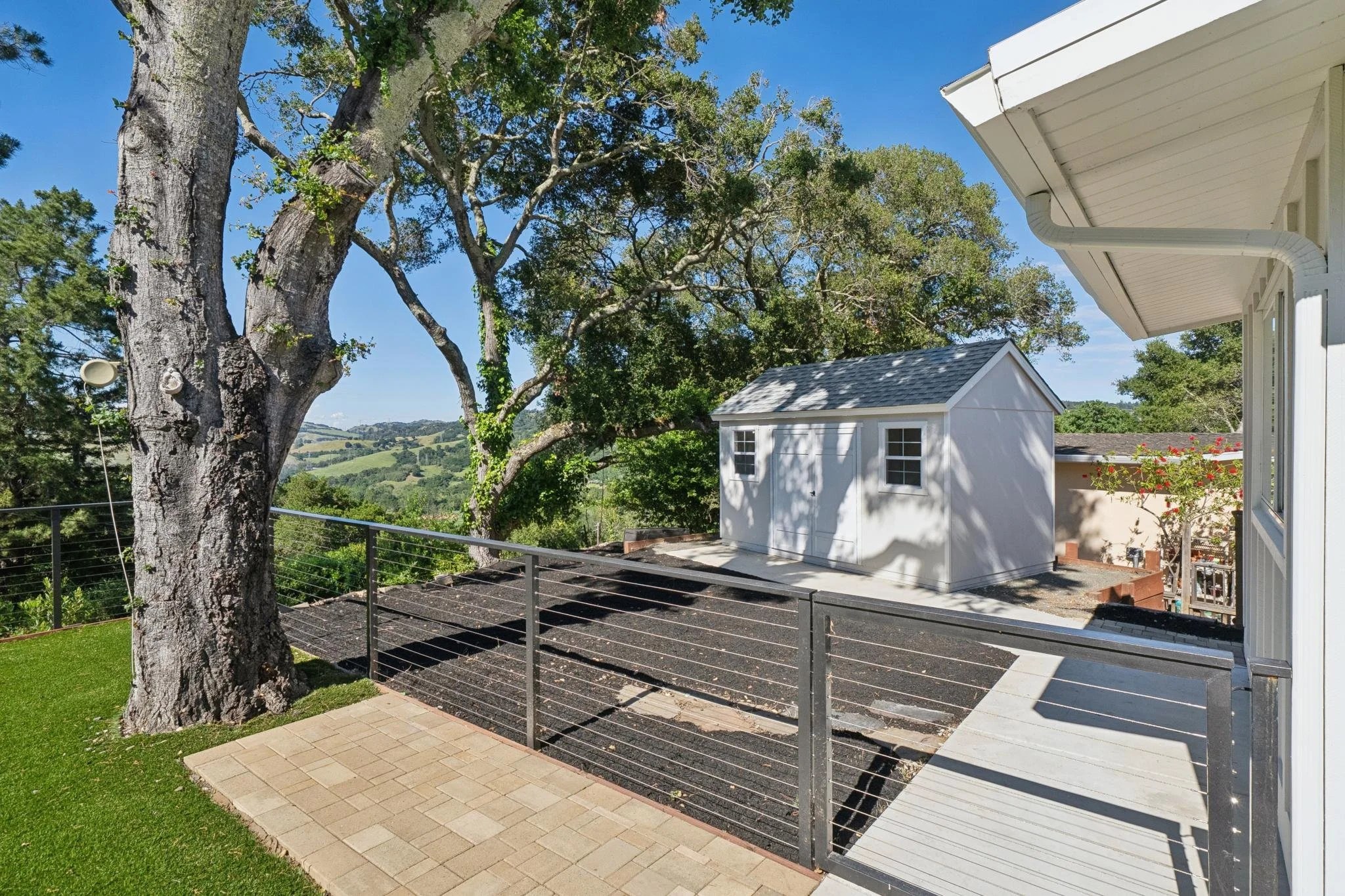 View of a backyard with a large tree, a white shed, a black metal railing, a patio area with paving stones, and a scenic landscape of rolling hills and greenery in the distance under a blue sky.