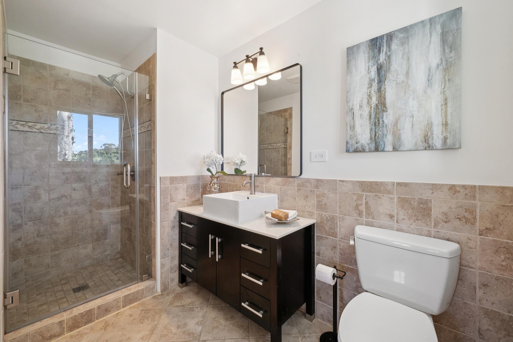 Modern bathroom with walk-in shower, white sink on dark vanity, toilet, mirror, abstract wall art, and decorative plants