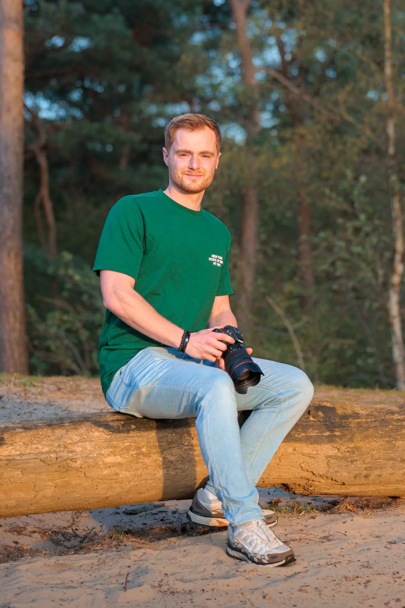 Een man met lichtbruin haar en een baard, zittend op een boomstam in een bos, terwijl hij een camera vasthoudt. Hij draagt een groene T-shirt, lichte spijkerbroek en witte sneakers. Het is zonnig en de achtergrond bestaat uit bomen en bladeren.