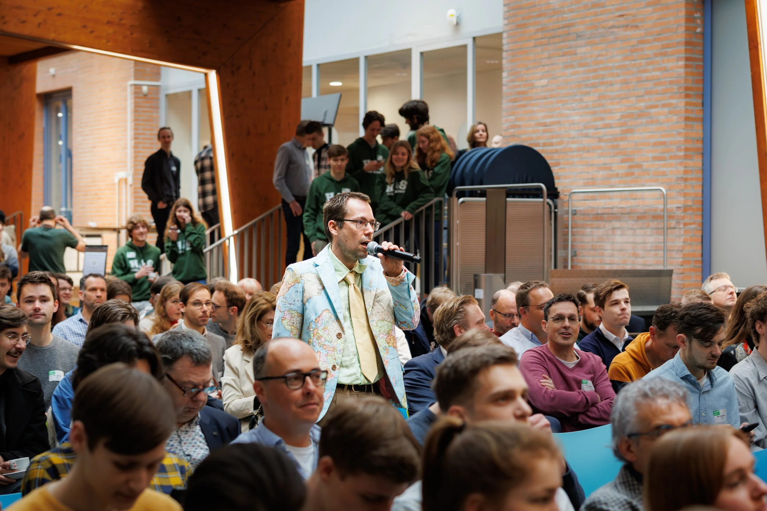 Een man met een wereldbol op zijn jas staat in een zaal met veel mensen tijdens een bijeenkomst, met een podium en een groep mensen op de achtergrond.