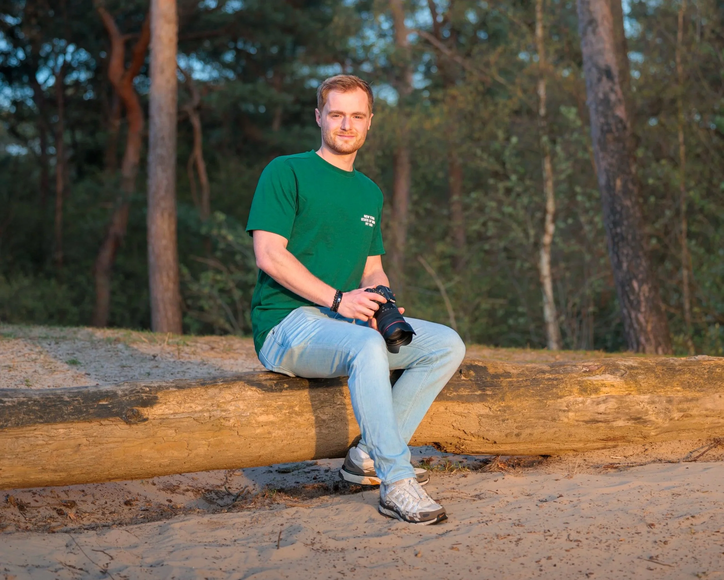 Een jonge man met een rode baard en haar, zittend op een grote stam in het bos, terwijl hij een camera vasthoudt. Hij draagt een groene T-shirt en lichte jeans, met de zonsondergang op de achtergrond.