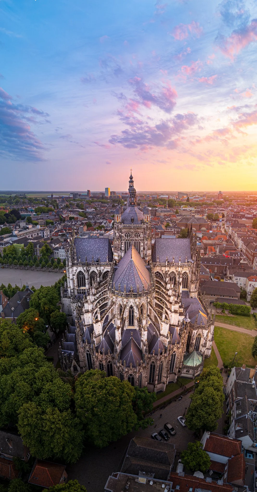 Een grote gotische kerk met hoge torens en gedetailleerde glas-in-loodramen, omgeven door bomen en een stadscentrum met gebouwen, onder een kleurrijke schemeringslucht.