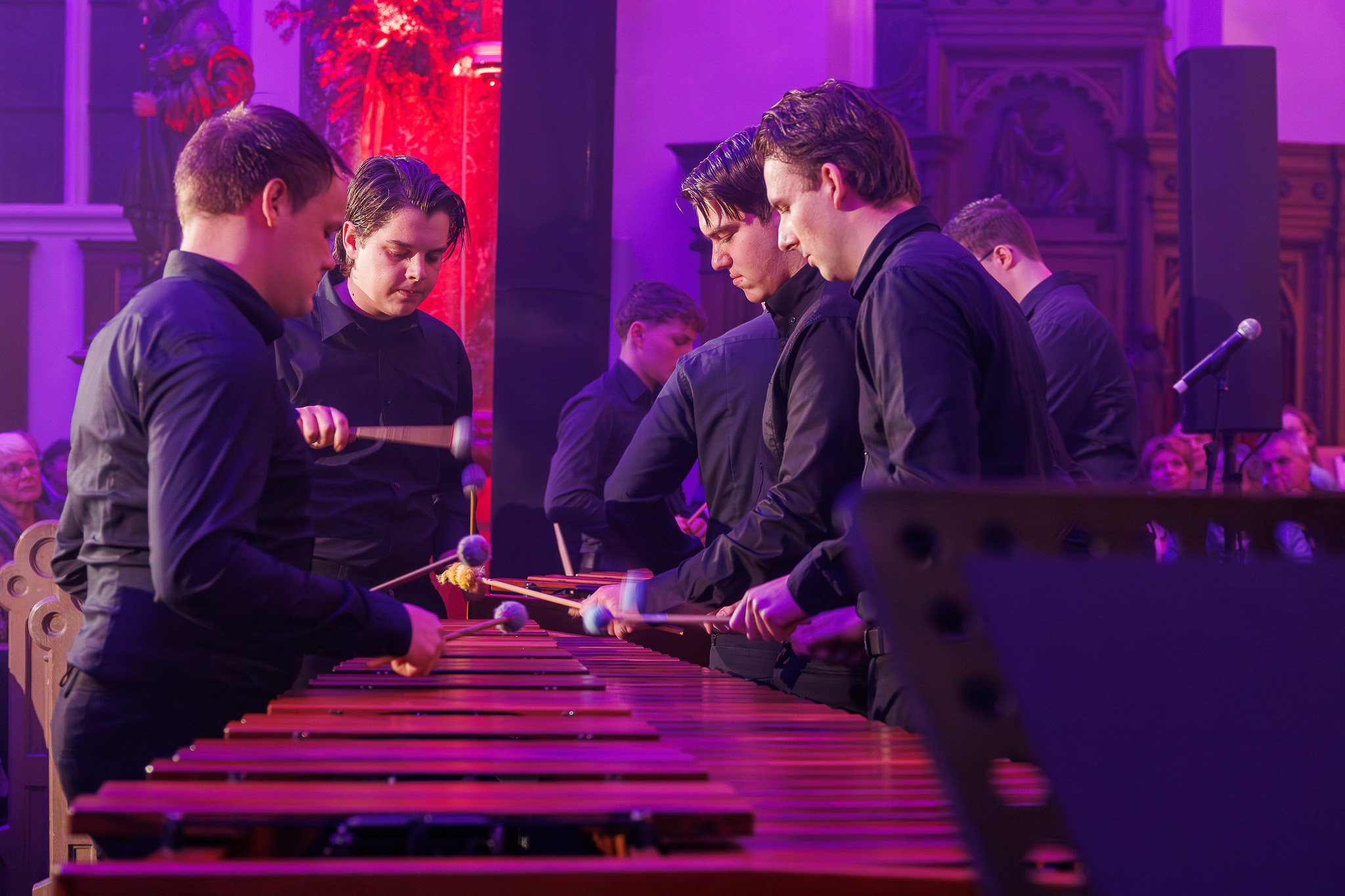 Vijf jonge mannen in zwarte kleding spelen op een grote houten xylofoon tijdens een concert in een kerk, met een publiek op de achtergrond.