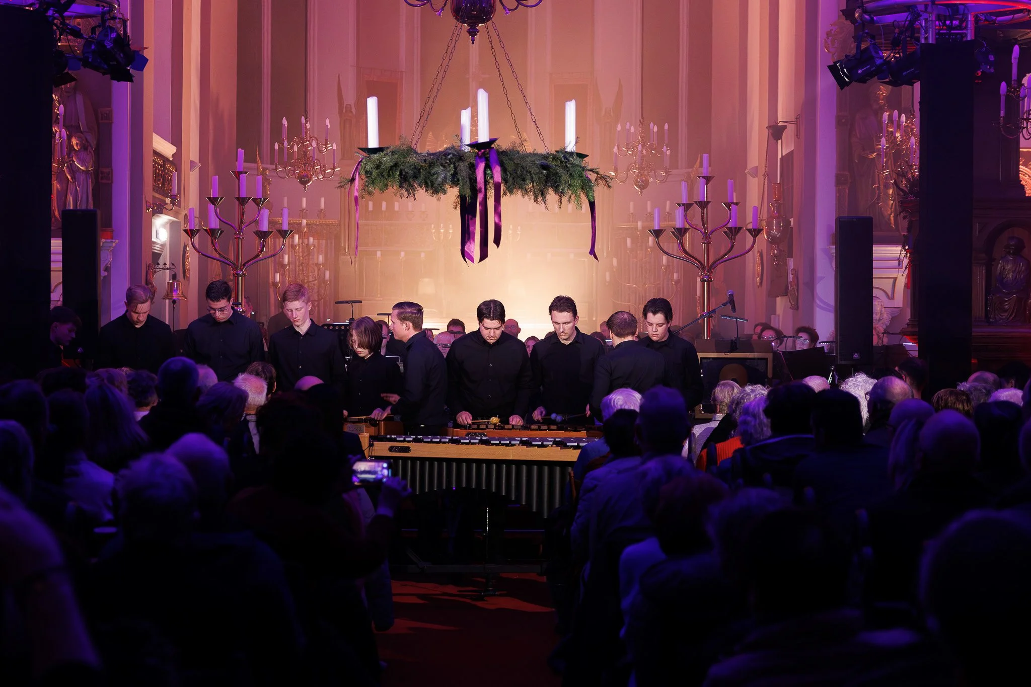 Mensen die een muziekuitvoering of orkest in een kerk of concertzaal bijwonen, met een decorisch interieur en grote kandelaars, onder een grote kroonluchter en kleurrijk verlichte ambiance.