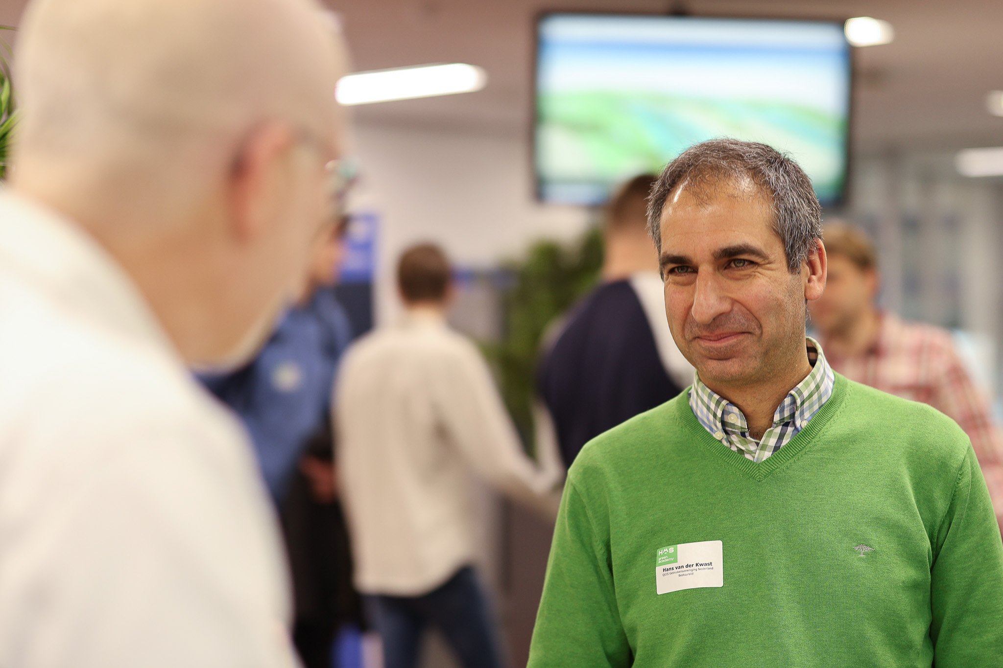 Man in groene trui met naamkaartje voor een conferentie, praat met iemand in een drukke, moderne kantooromgeving.