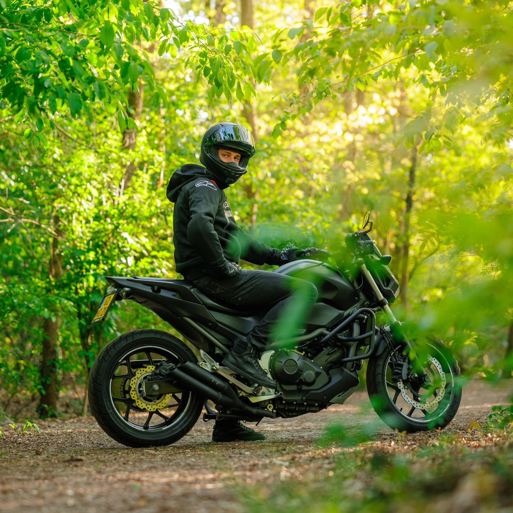 Een persoon in motorkleding en helm die op een zwarte motorfiets in een bosrijk gebied rijdt.