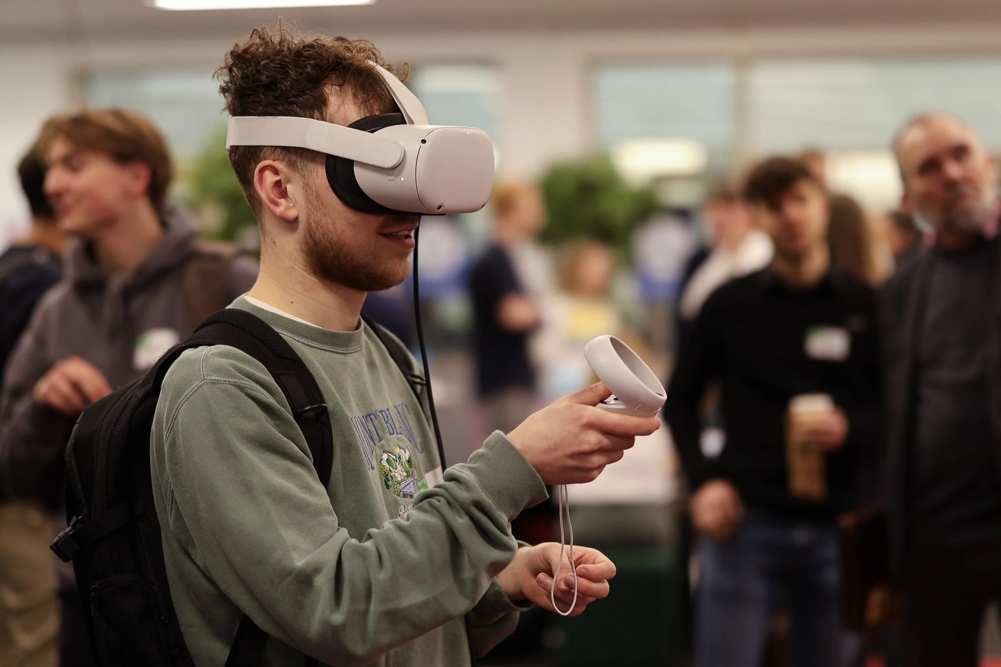 Man met een virtual reality-headset die een controller vasthoudt, in een drukke ruimte met meerdere mensen.