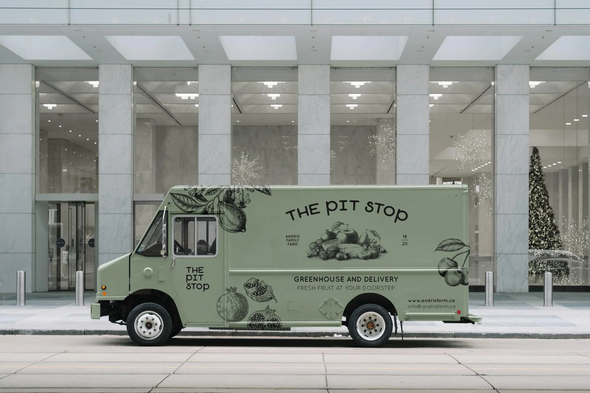 A green fruit delivery truck parked in front of a modern building with glass windows and a Christmas tree inside, promoting 'The Pit Stop' farm-to-doorstep produce service.