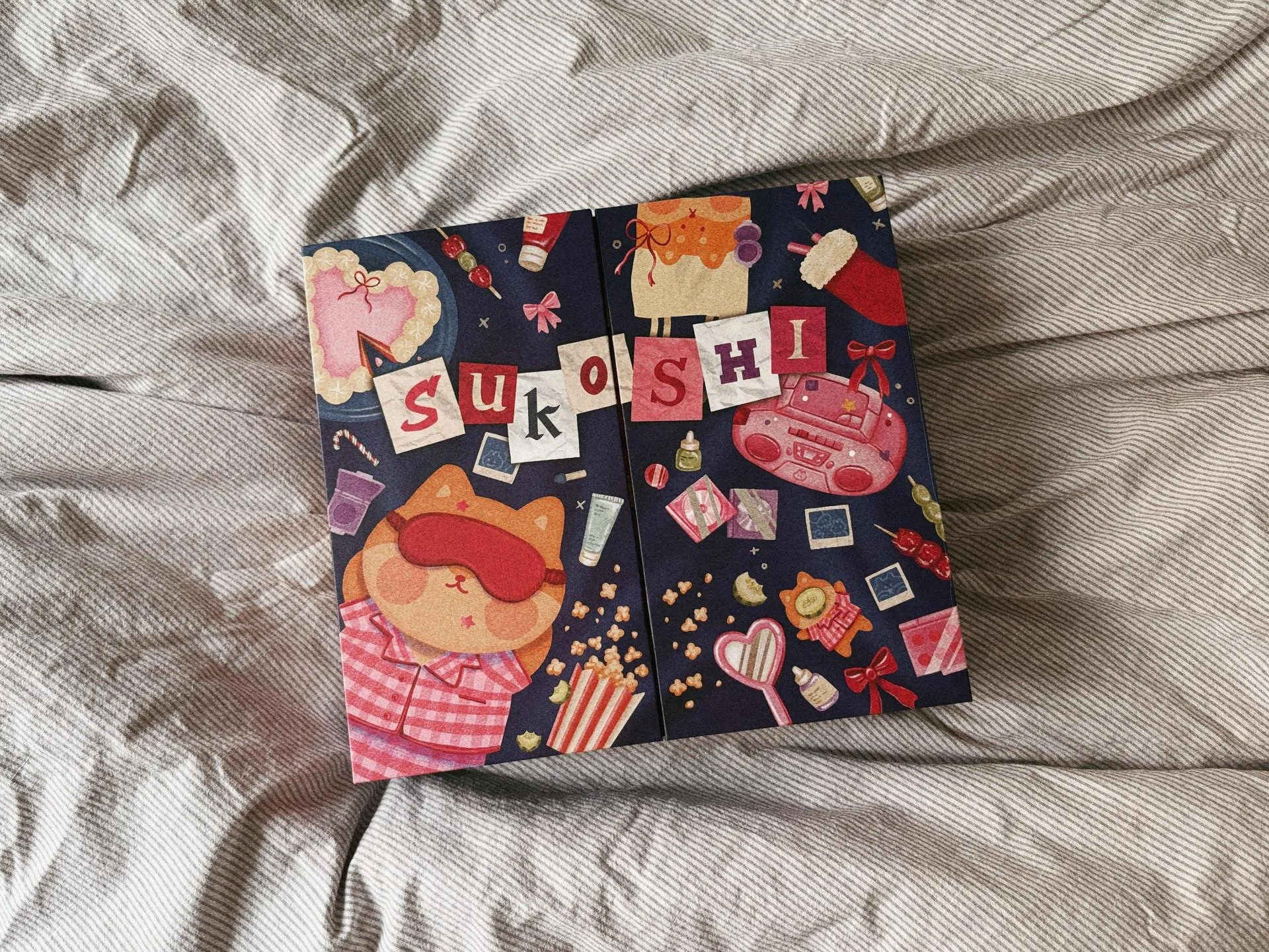 Colorful greeting card on a striped fabric surface, with illustrations of a popcorn box, a bear with a sleep mask, a radio, sweets, and the word 'SUSHI' spelled out with letter blocks.