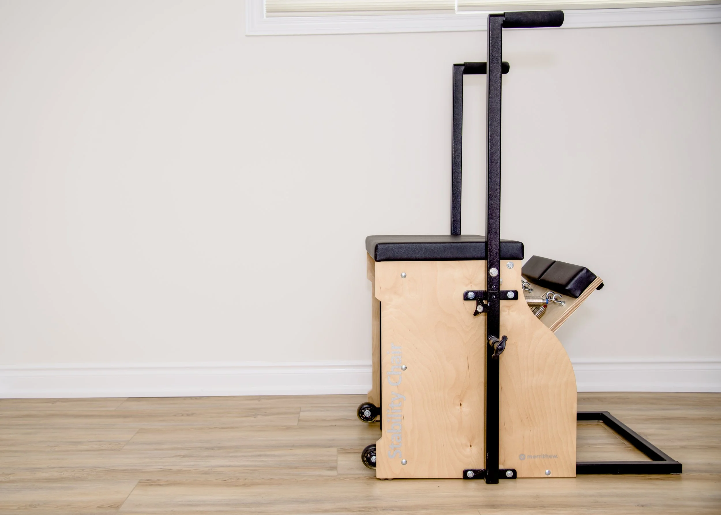 A wooden chair with black padding and metal frame, designed for physical therapy or exercise, positioned against a plain white wall on a light wood floor.