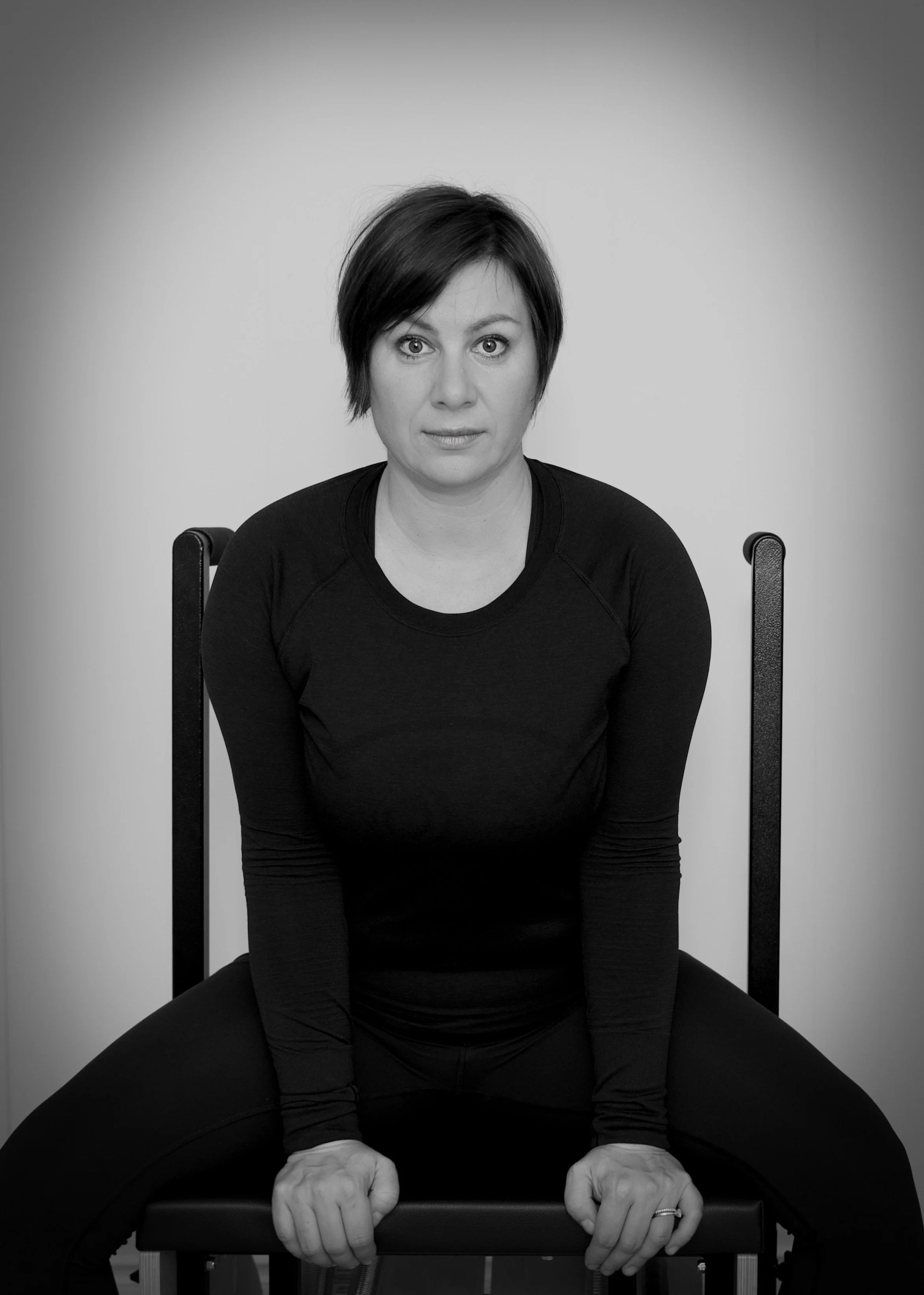 A woman with short dark hair, wearing a black long-sleeve shirt, sitting on a chair with her hands gripping the edge of a table, looking directly at the camera against a plain background.