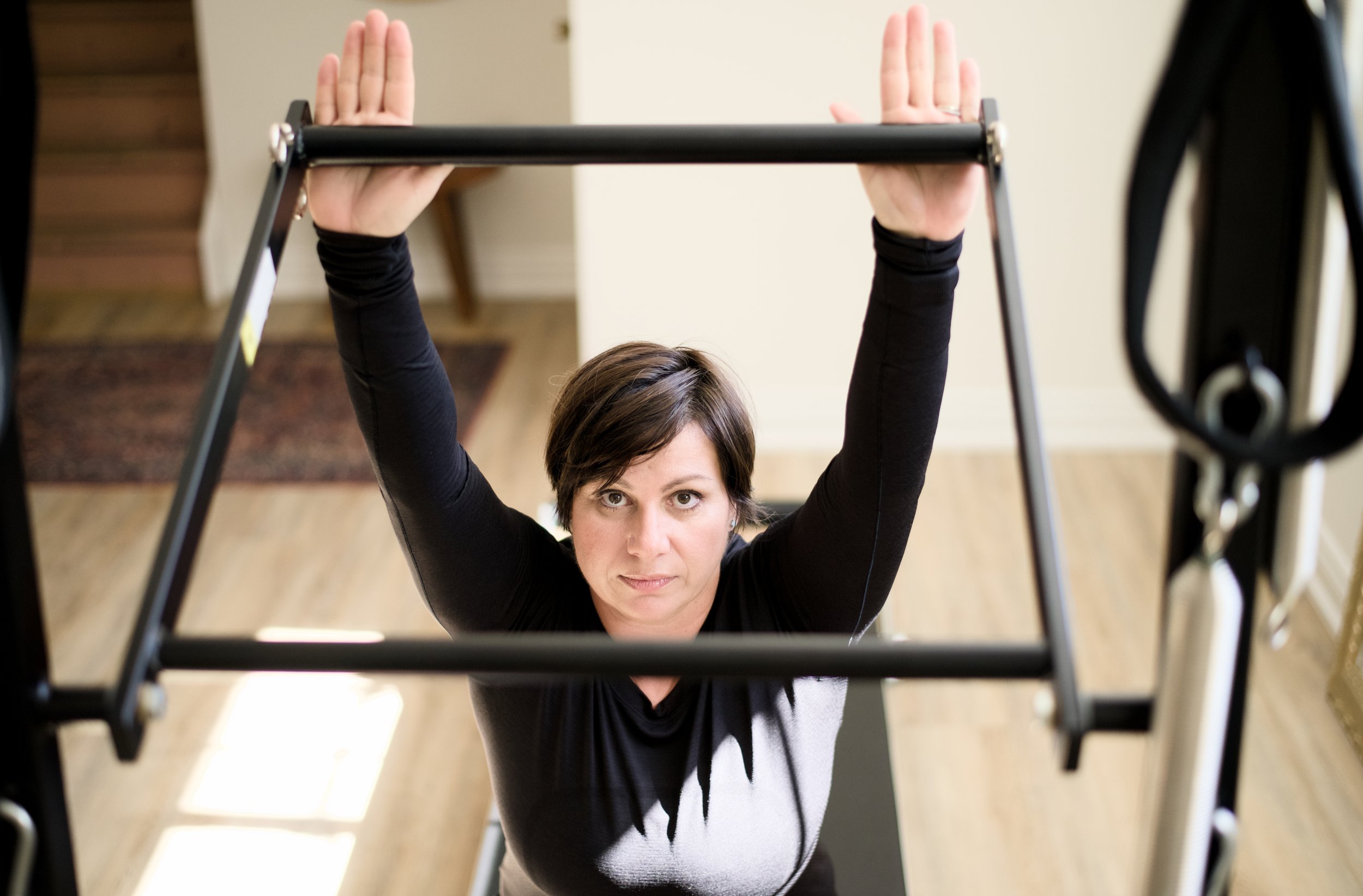 A woman in black long sleeve shirt doing a Pilates exercise using a reformer machine in a home setting with hardwood floors.
