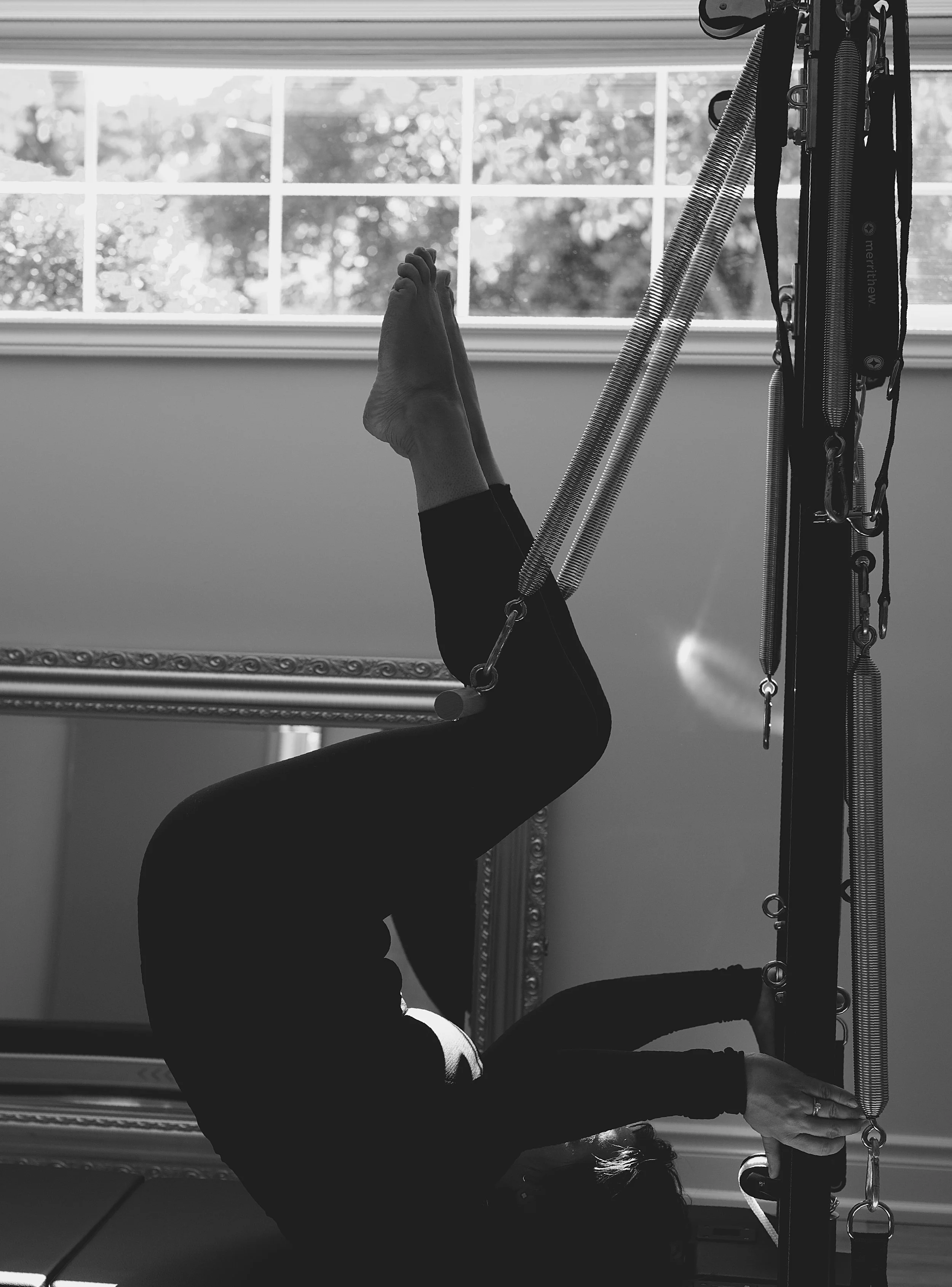 A person practicing aerial Pilates, hanging upside down using straps attached to a wall or ceiling, with a window in the background and sunlight coming through.