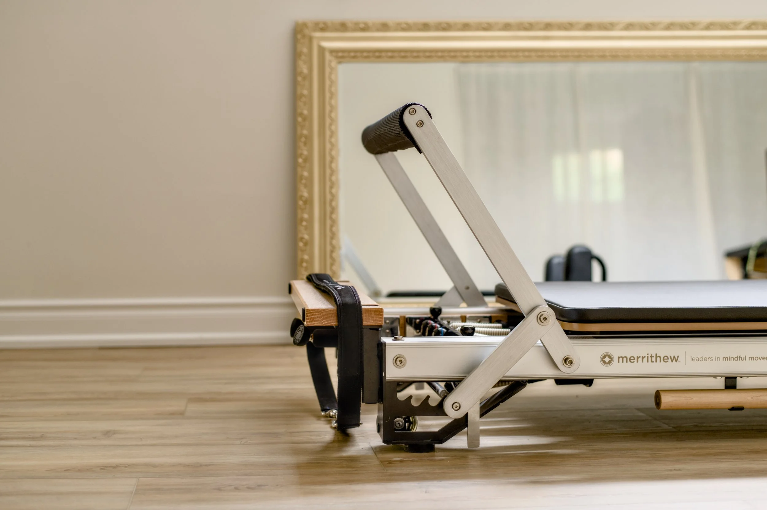 A portable massage or therapy chair with an adjustable backrest, placed on a wooden floor in front of a large ornate mirror. The chair has a metal frame and is branded with 'merrithew.'