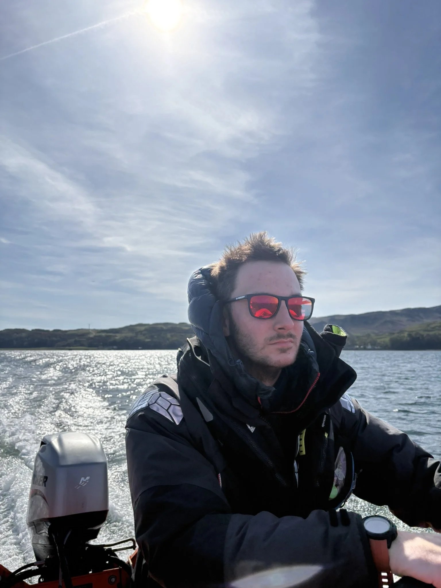 A man with sunglasses, wearing a jacket and a hoodie, is on a boat with a motor, riding on a body of water. The background includes hills and a partly cloudy sky with the sun shining.