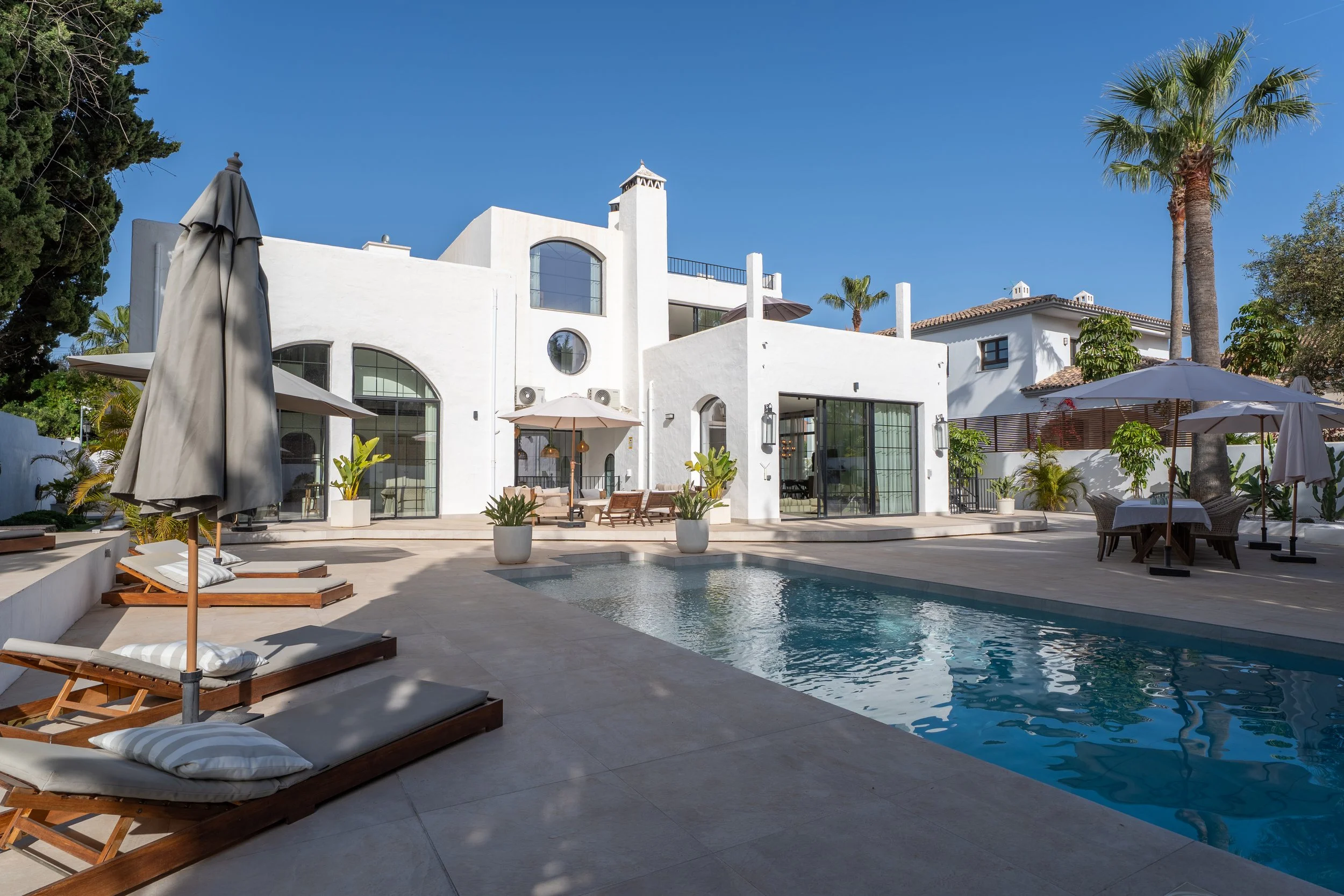 A luxurious white house with a swimming pool in the foreground, surrounded by lounge chairs, umbrellas, and palm trees, under a clear blue sky.